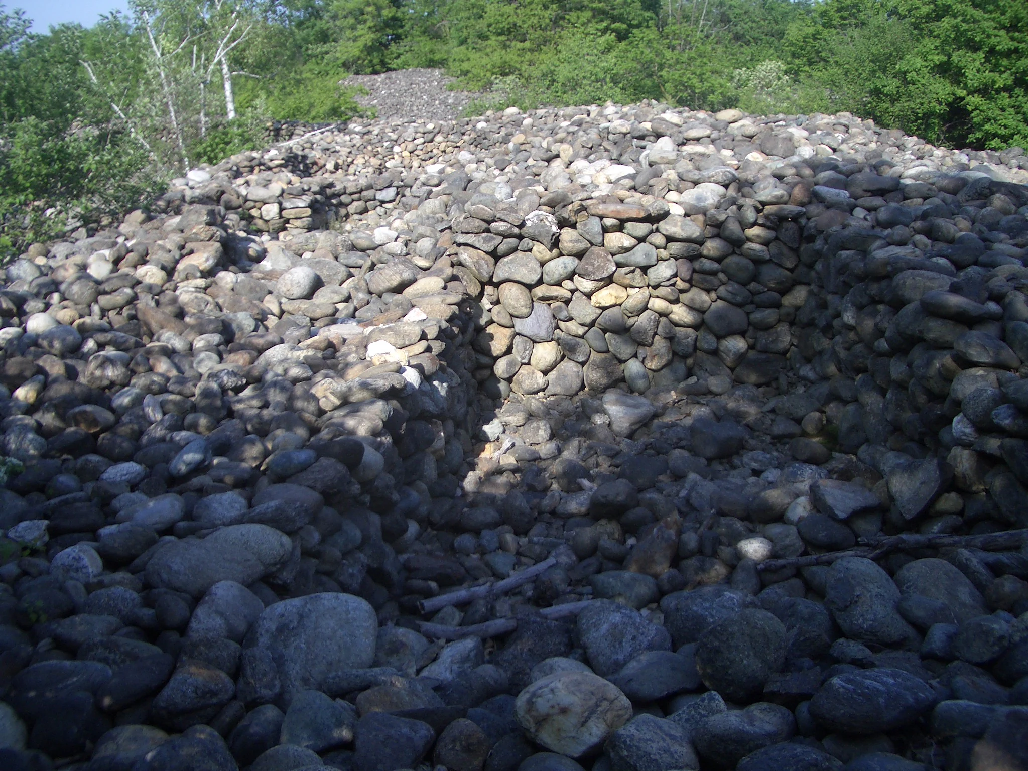 A close up view of ancient stone walls and rubble composed of round, grey river stones, set among green trees.