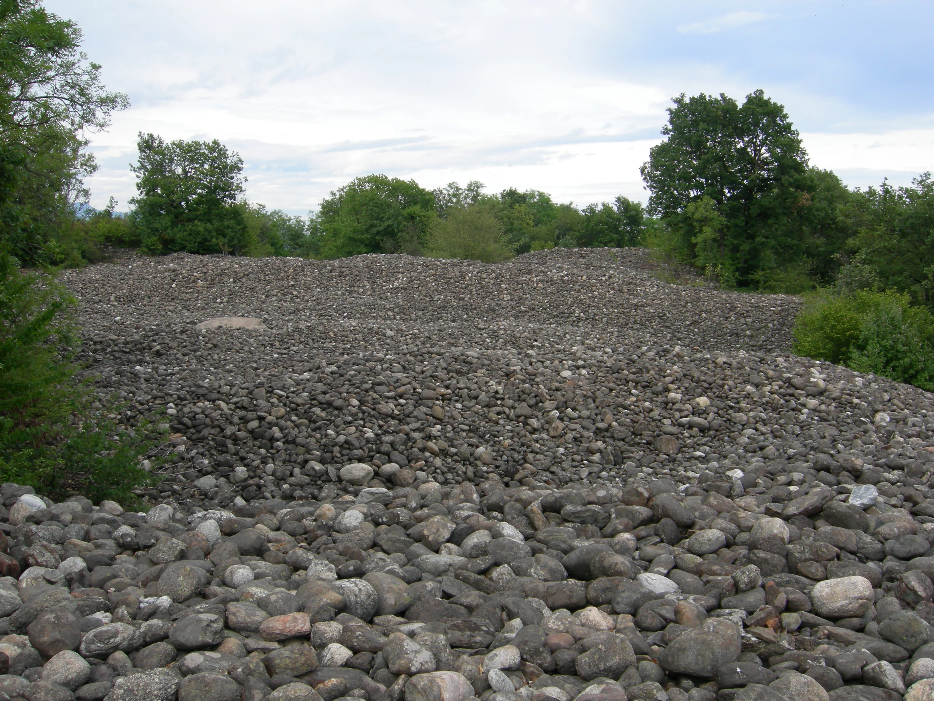 A vast, slightly hilly field covered in grey rounded river stones, bordered by green deciduous trees under a cloudy sky.