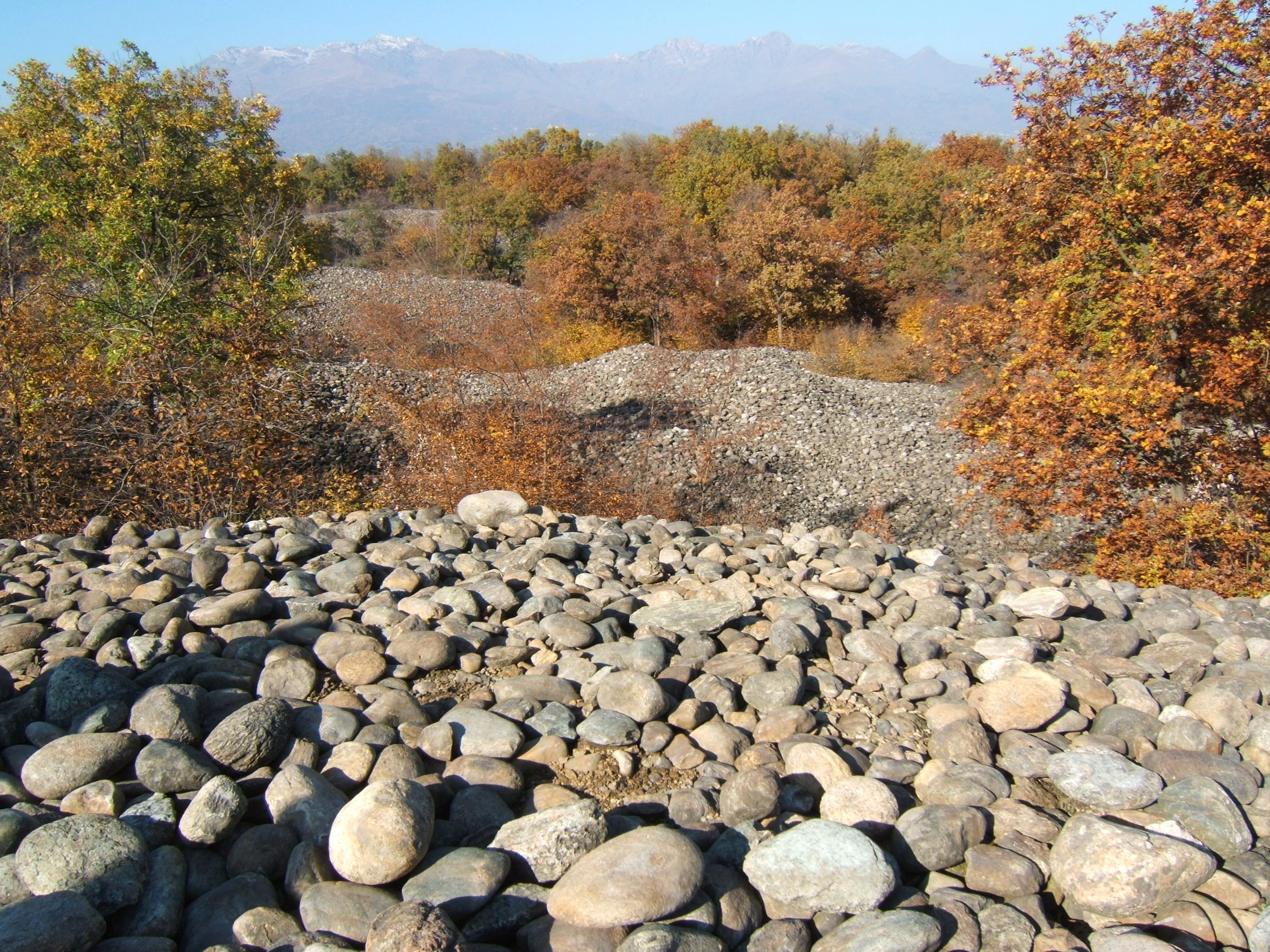 A landscape view featuring piles of rounded grey river stones covering the ground, surrounded by trees with autumn foliage and distant mountains under a clear sky.