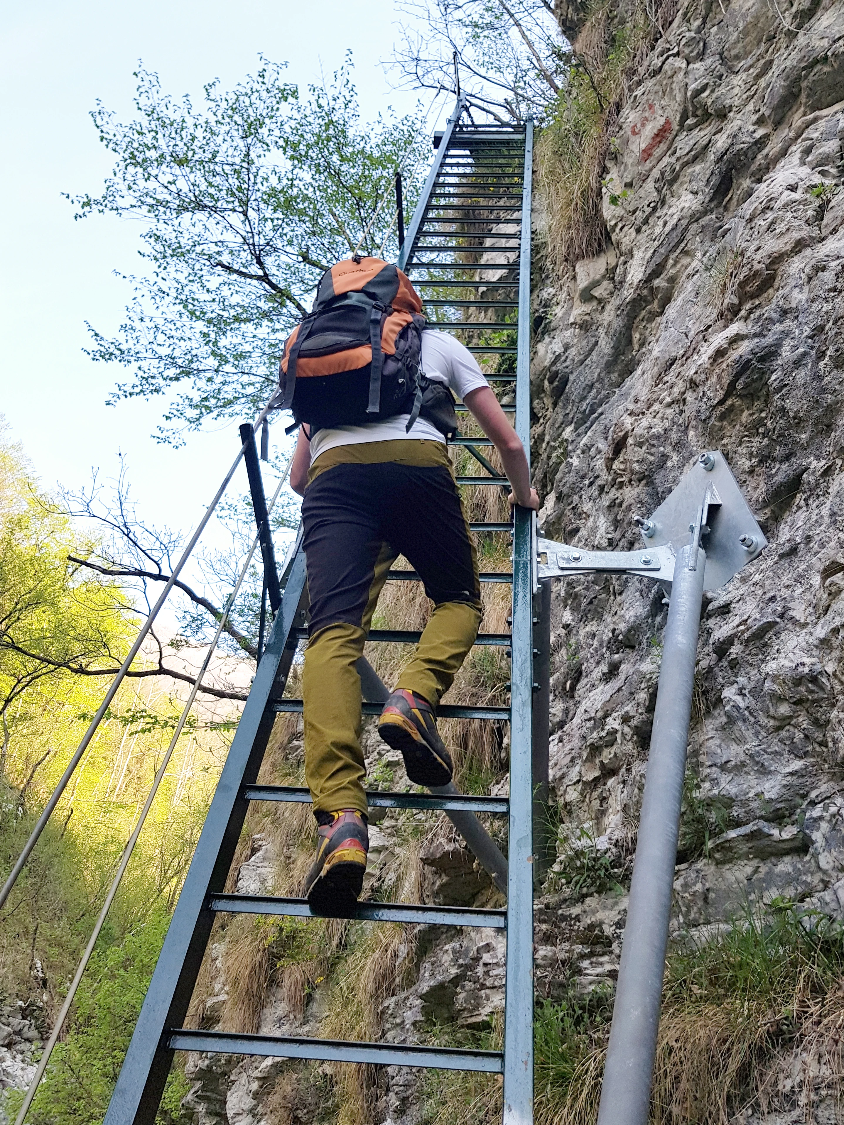Un excursionista con mochila subiendo por una escalera metálica estrecha y empinada fijada a un acantilado rocoso en un sendero.