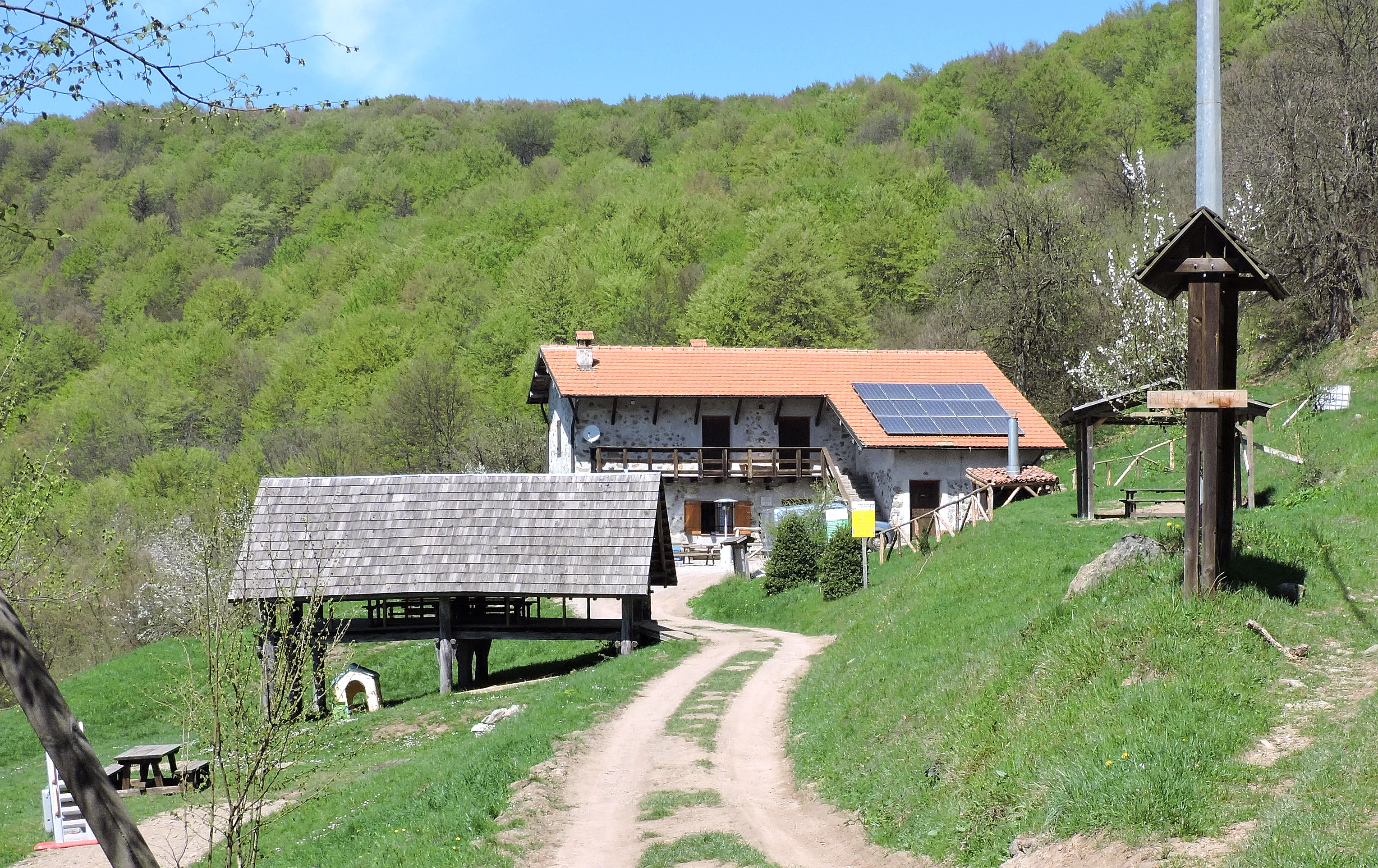 A stone farmhouse with a terracotta roof, solar panels, and an adjacent open-sided wooden structure, nestled in a green valley in the Adelasia Nature Reserve.