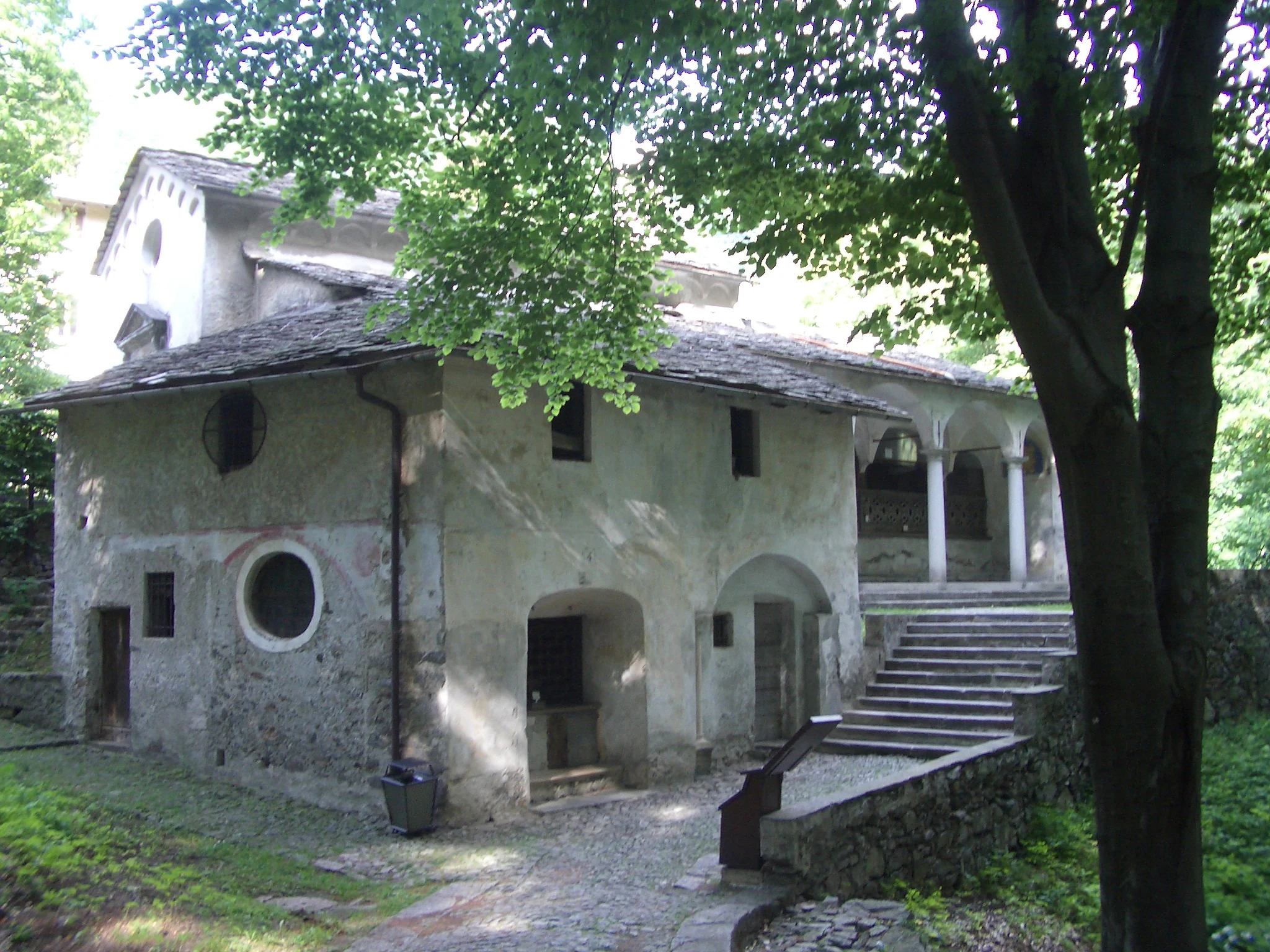 A stone chapel with a sloped roof, arched entrances, and a stone staircase, nestled in a forested setting.