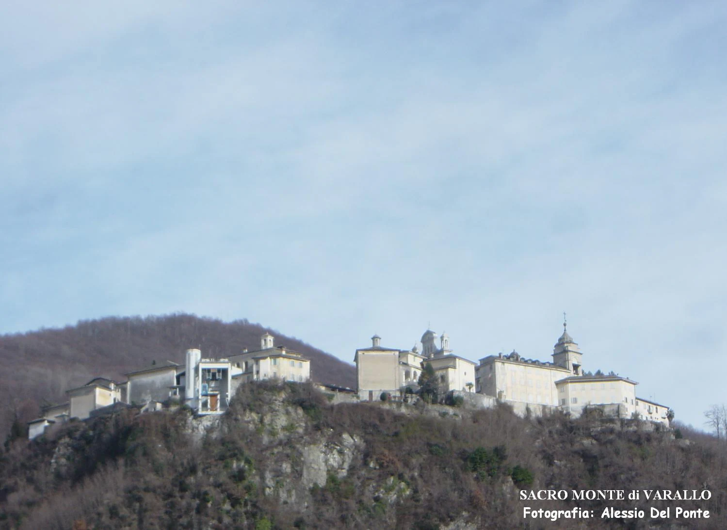 A group of historic stone chapels and buildings with tiled roofs atop a forested rocky hill under a bright, overcast sky.