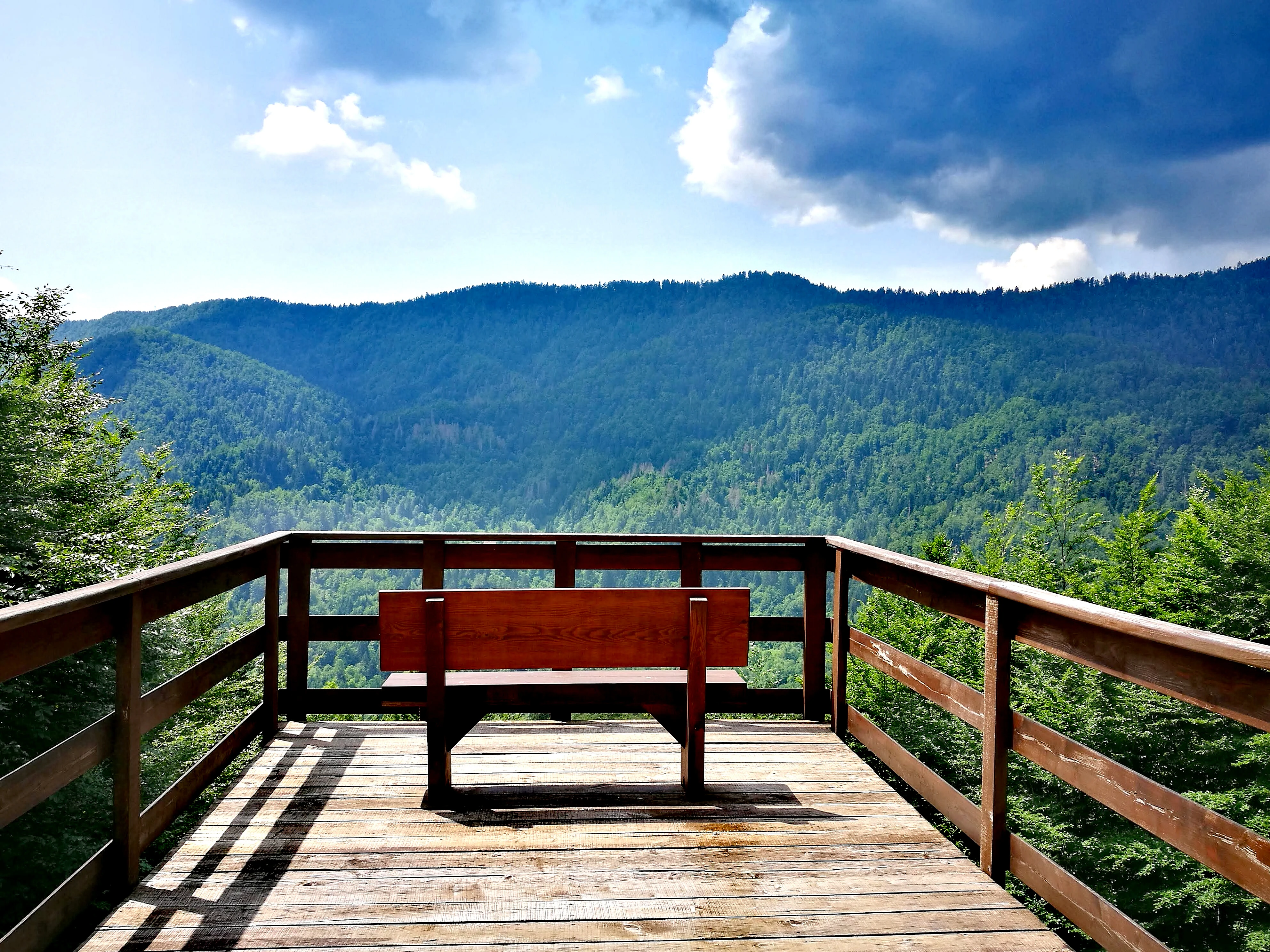 Wooden deck with brown bench, wooden railings, and expansive forested mountain range under partly cloudy sky