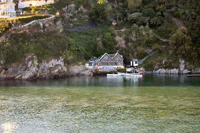 Una pequeña barcaza de ferry atracada en un embarcadero de piedra a orillas del río Gannel, con acantilados boscosos y un edificio visibles al fondo.
