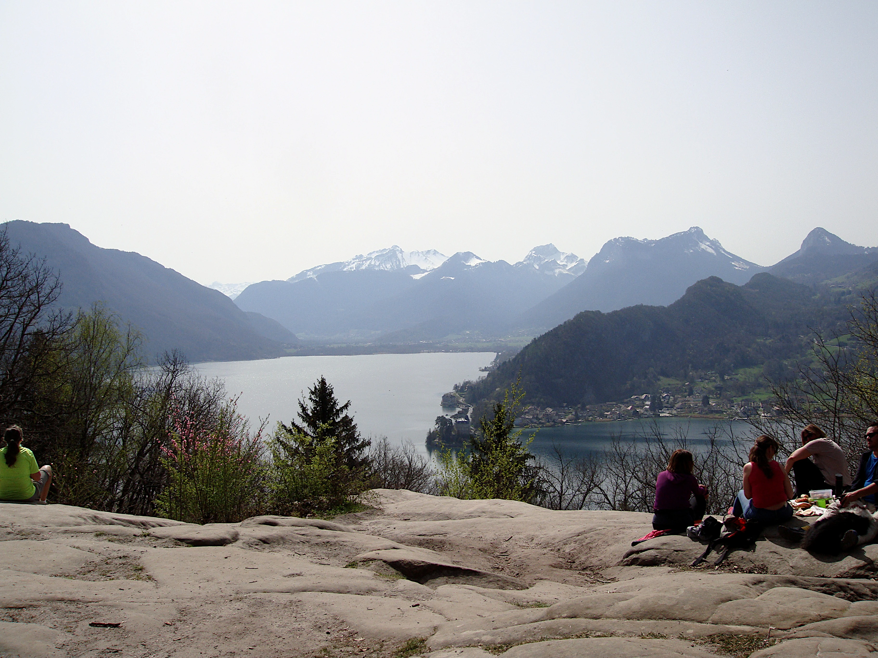 Una vista de gran angular desde una colina rocosa con vistas a un gran lago rodeado de montañas, con varios excursionistas descansando en las rocas en primer plano.