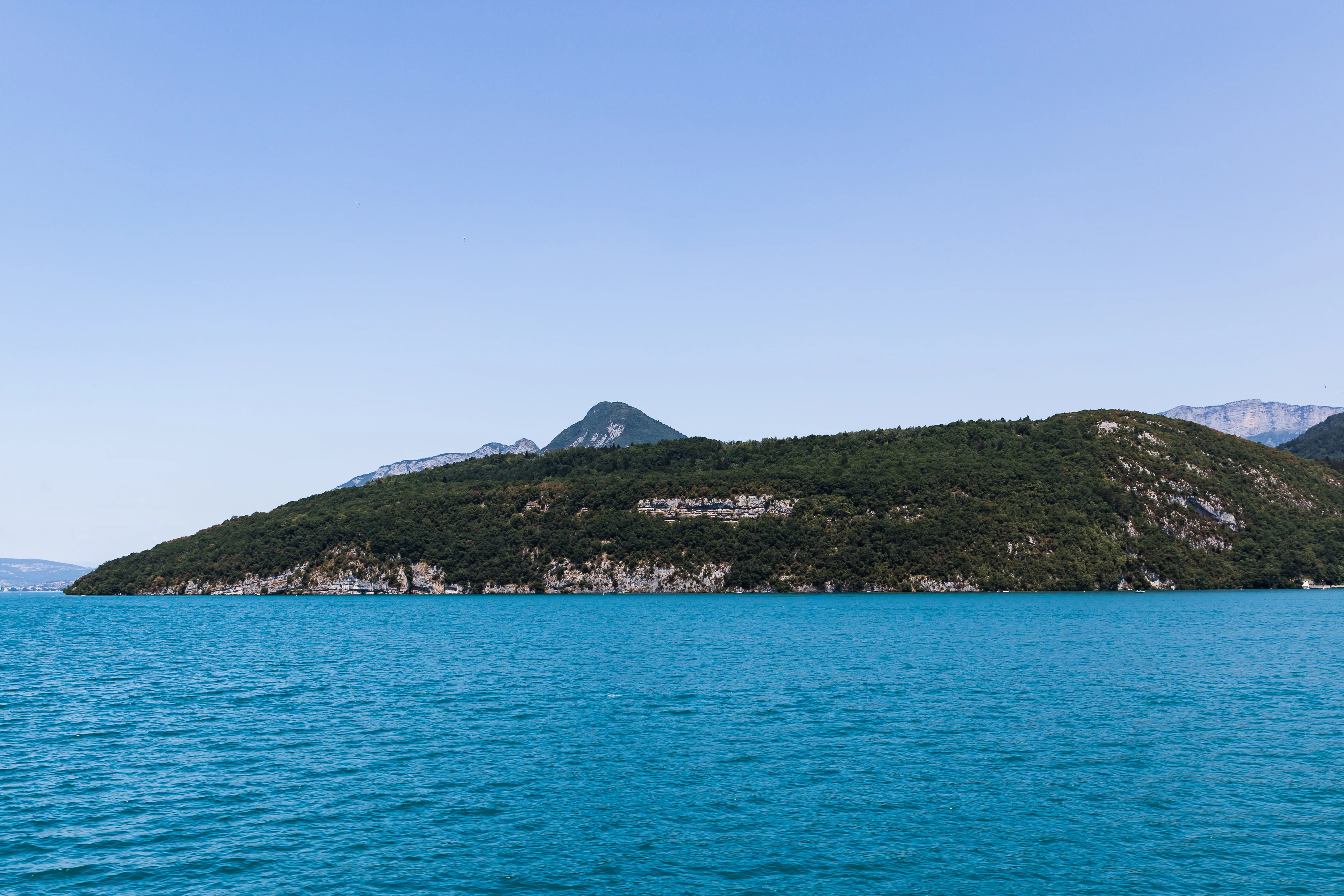 Una amplia vista panorámica sobre las aguas azul brillante del lago de Annecy hacia las laderas meridionales boscosas y rocosas del promontorio de Roc de Chère, con un pico más alto al fondo bajo un cielo despejado.