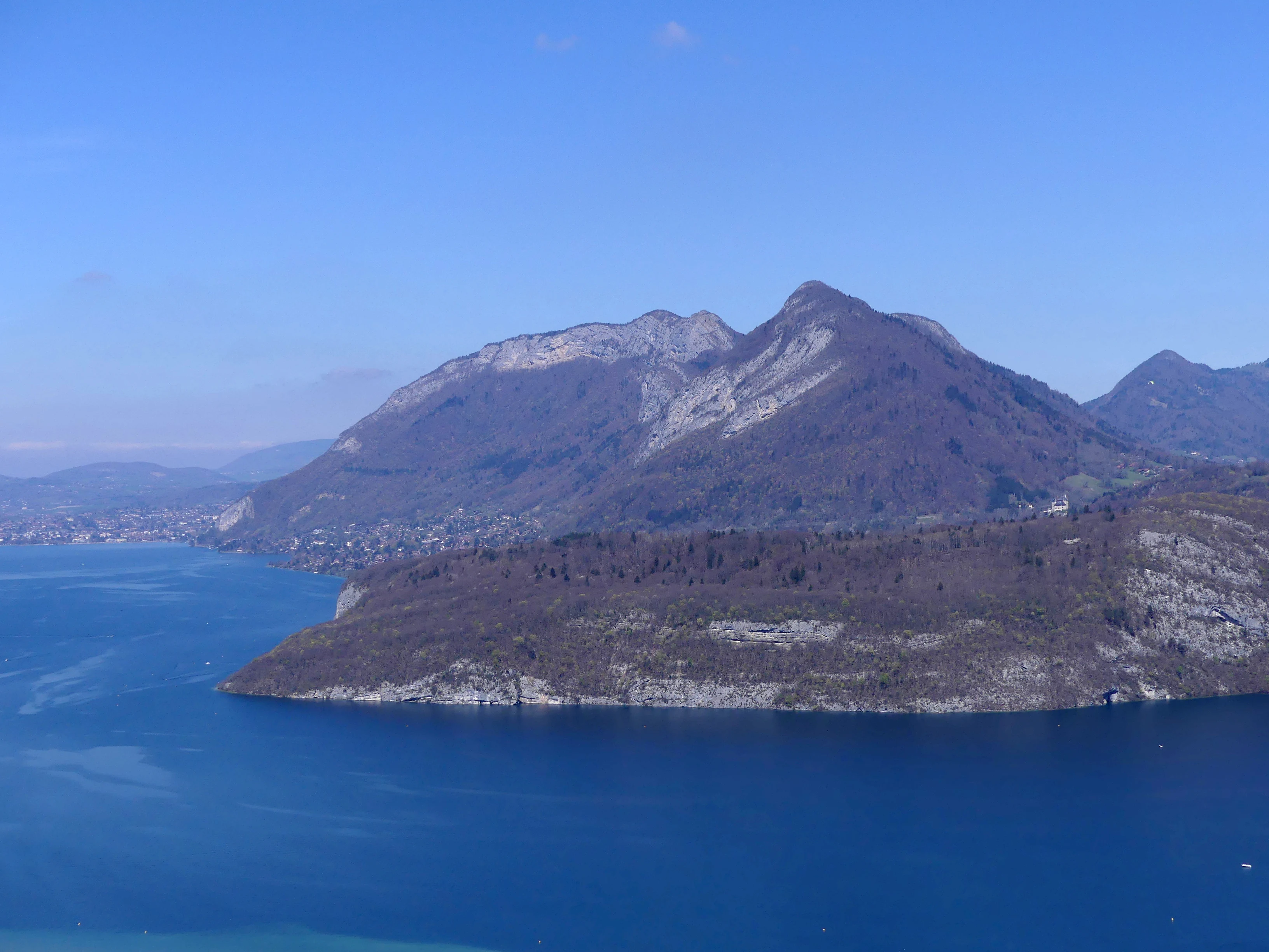 Una vista panorámica en ángulo alto que muestra un cabo rocoso y boscoso que se adentra en un gran lago azul, con montañas elevándose al fondo bajo un cielo despejado.