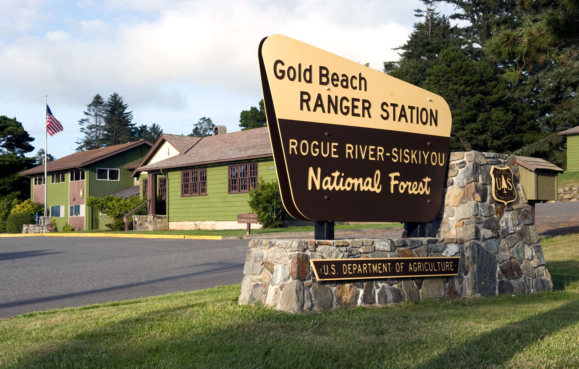 A large wooden park entrance sign reading Gold Beach Ranger Station and Rogue River-Siskiyou National Forest, mounted on a stone base, with green ranger station buildings in the background.