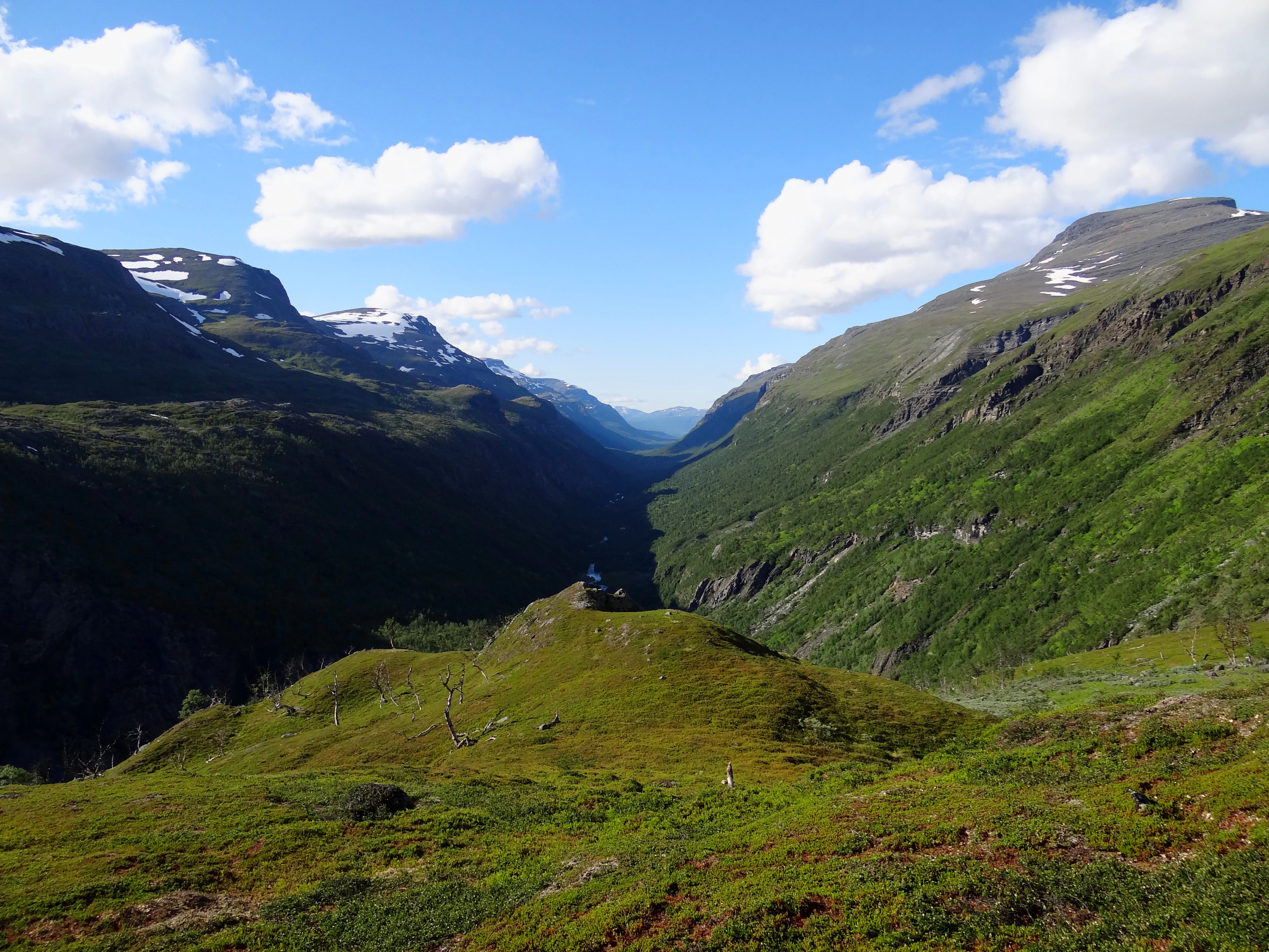 A wide valley with steep green slopes and snow-capped mountains in the distance under a blue sky with scattered clouds.