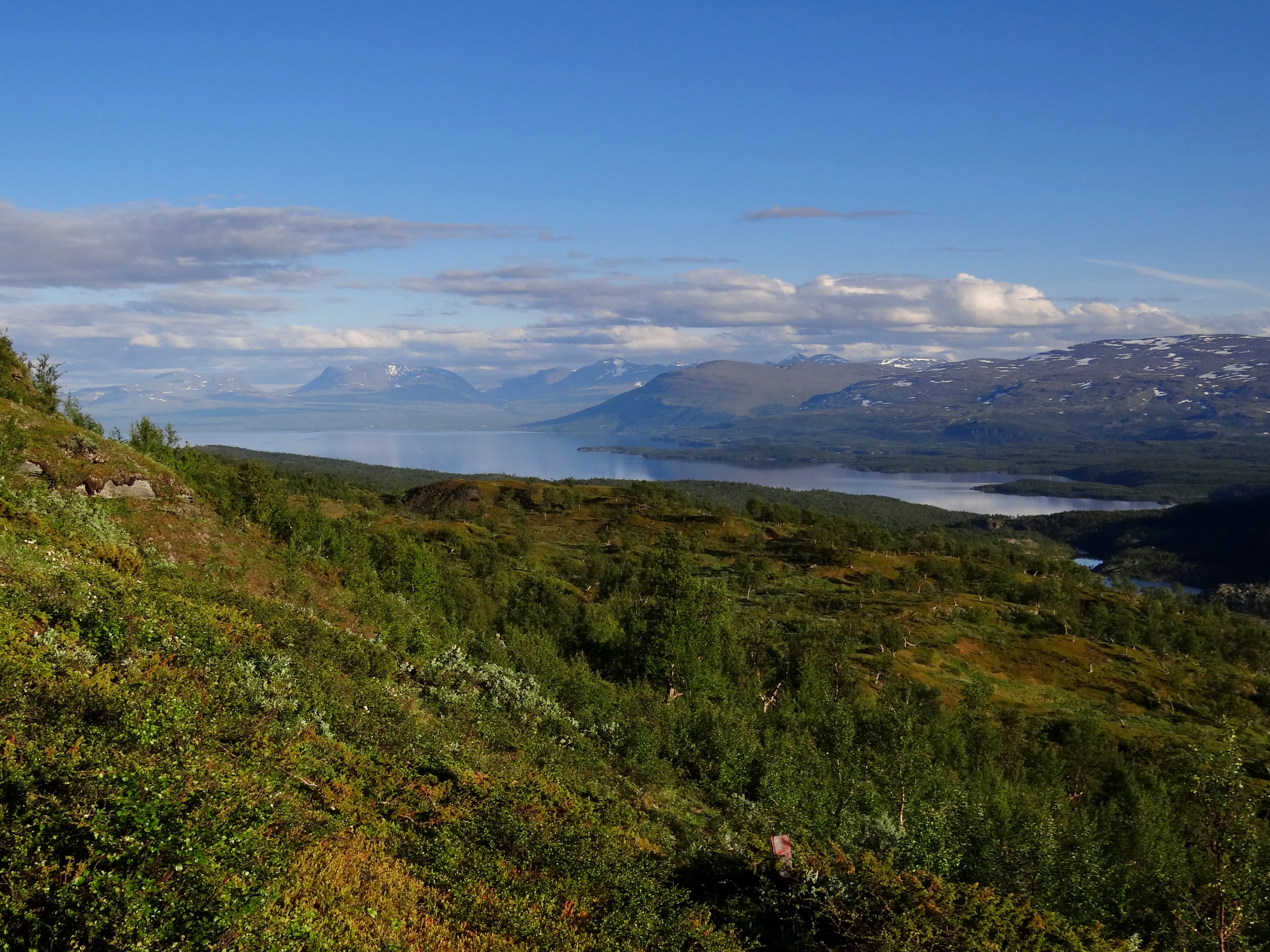 Panoramic landscape view showing a large lake surrounded by hills and mountains under a partly cloudy blue sky