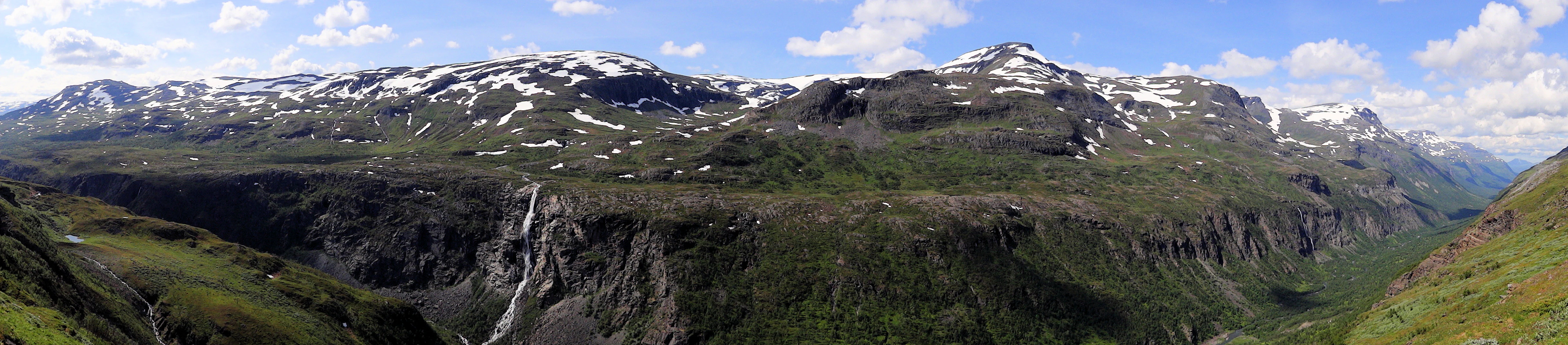 Panoramic view of Sørdalen canyon with multiple snow-capped mountains, green slopes, and a waterfall under a partly cloudy sky
