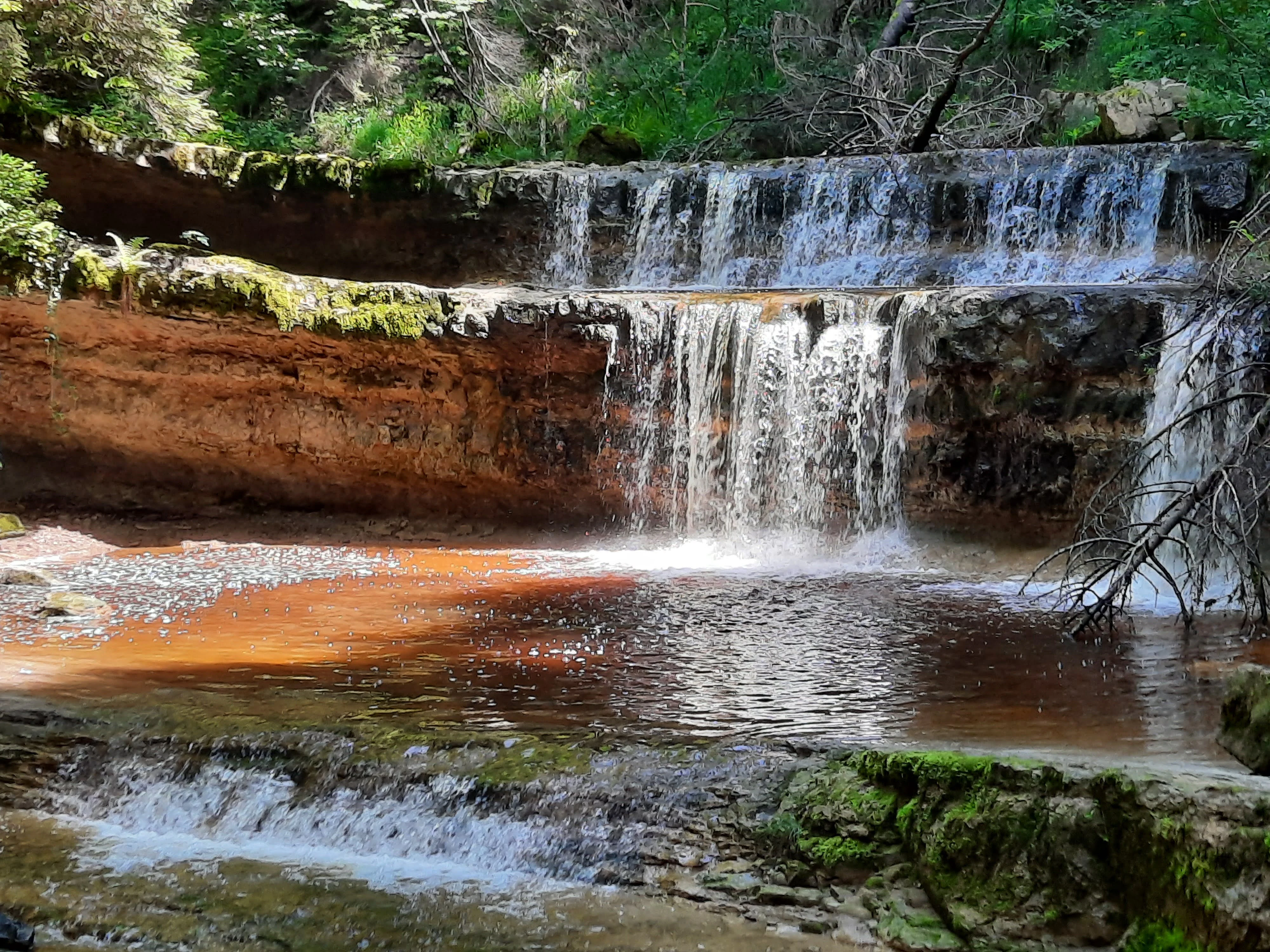 A multi-tiered waterfall cascading over reddish, layered rock formations into a calm, dark-bottomed pool, surrounded by lush green forest vegetation.