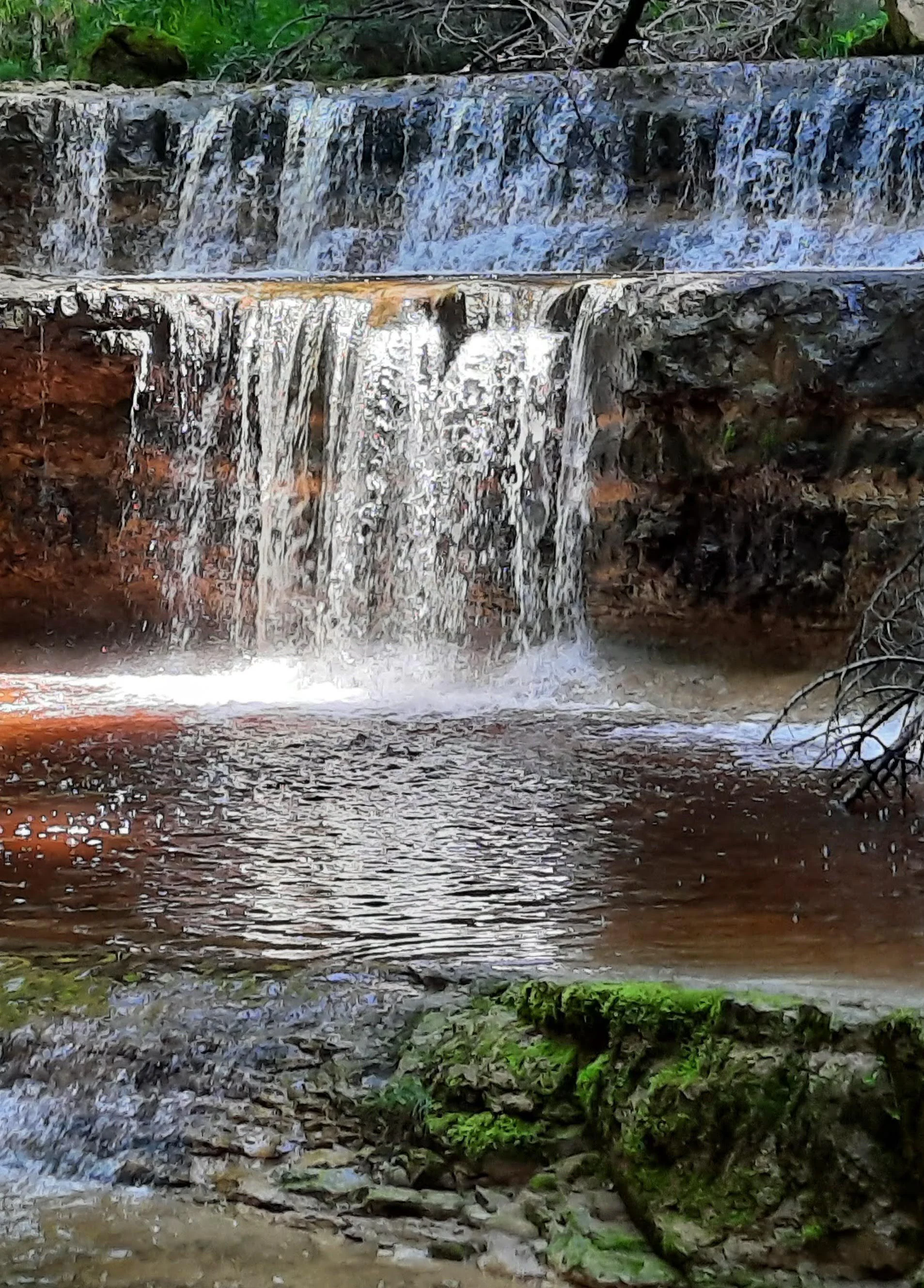 A small waterfall flowing over layered, reddish-brown rock formations into a calm, dark pool with mossy foreground rocks.