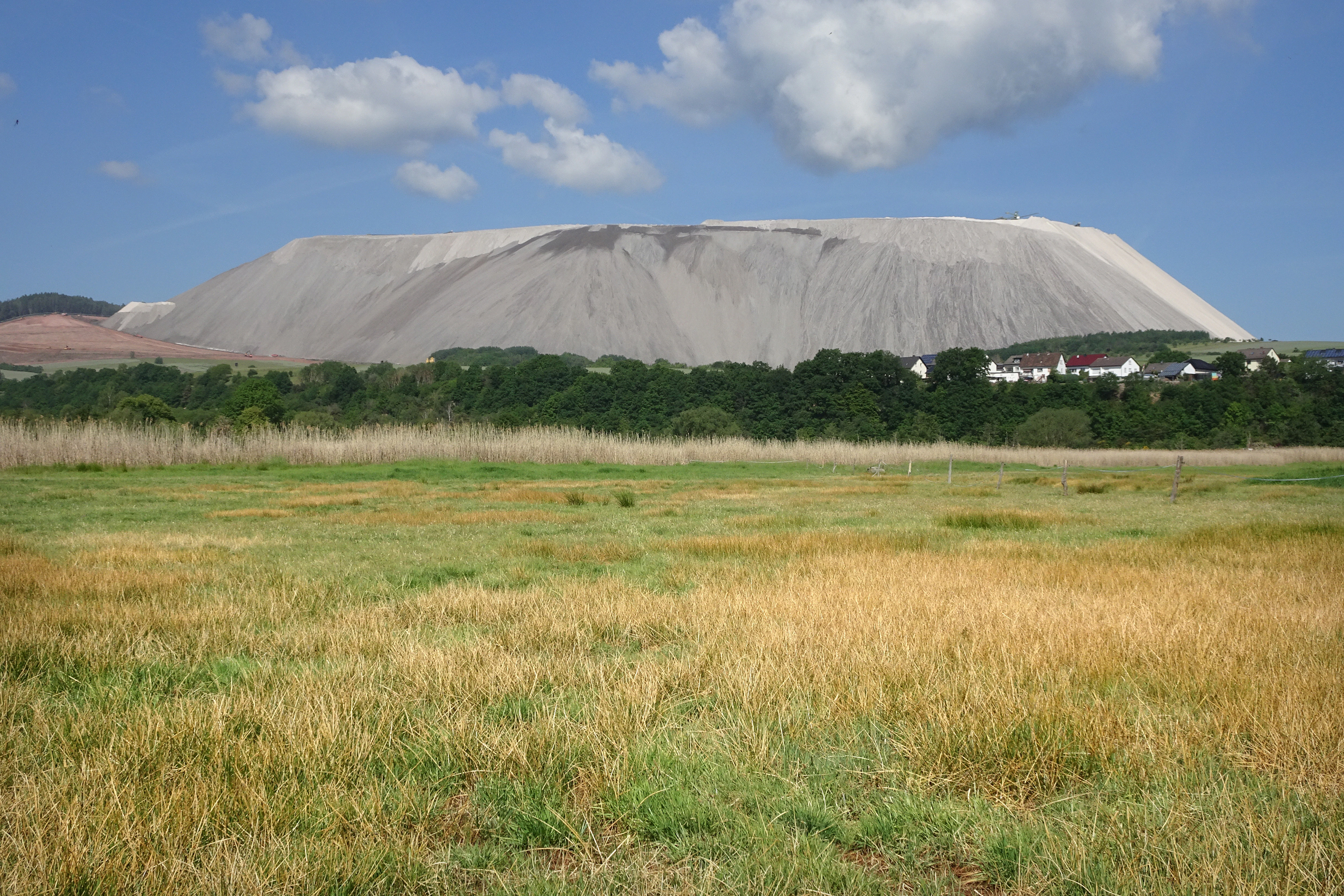 A wide view of a dry grassy meadow in the foreground leading to a line of trees and houses, with a massive gray, flat-topped artificial tailings hill rising in the distance under a blue sky.