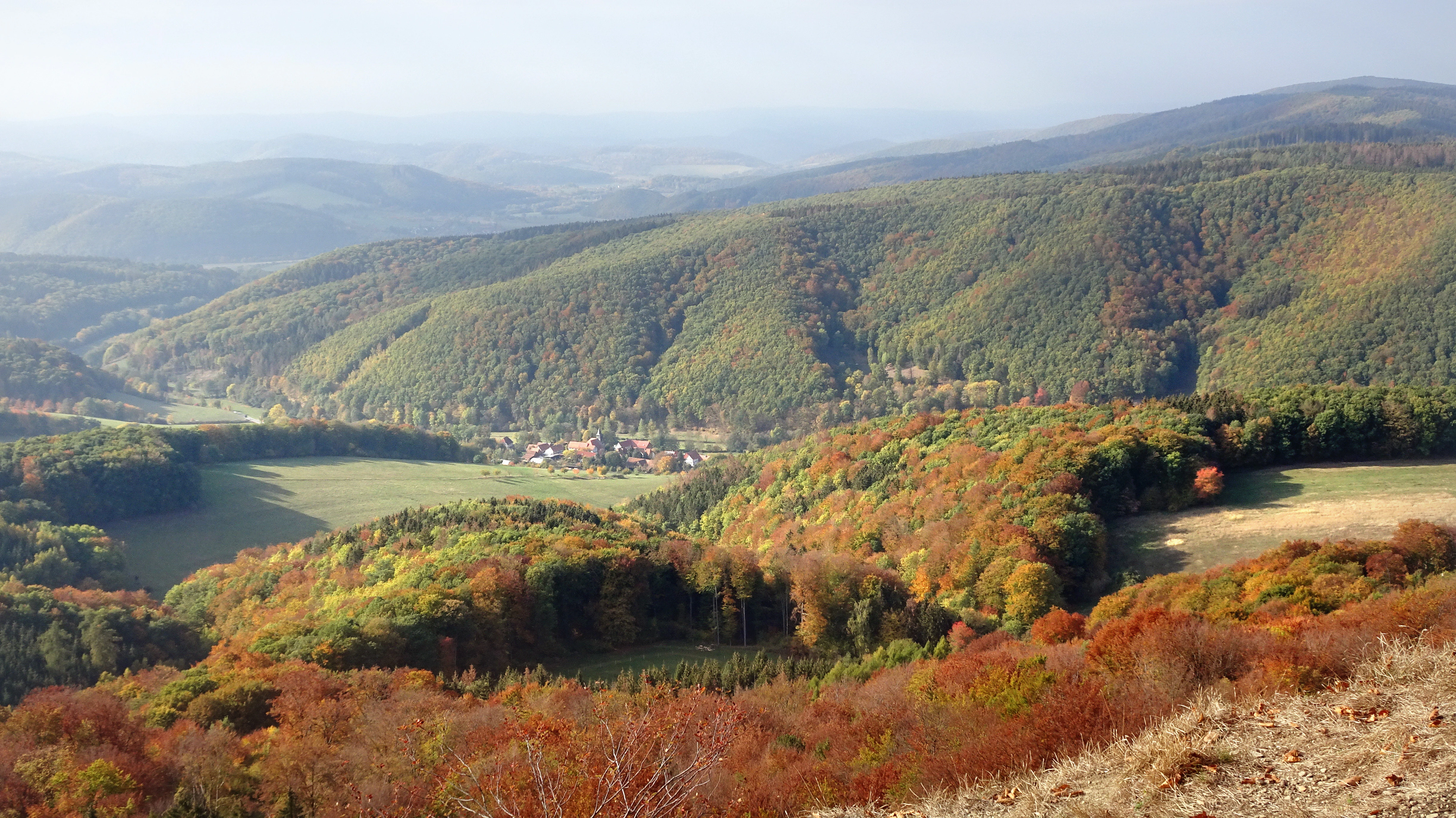 A wide aerial view of a valley with a small village surrounded by forested mountain slopes displaying autumn colors.