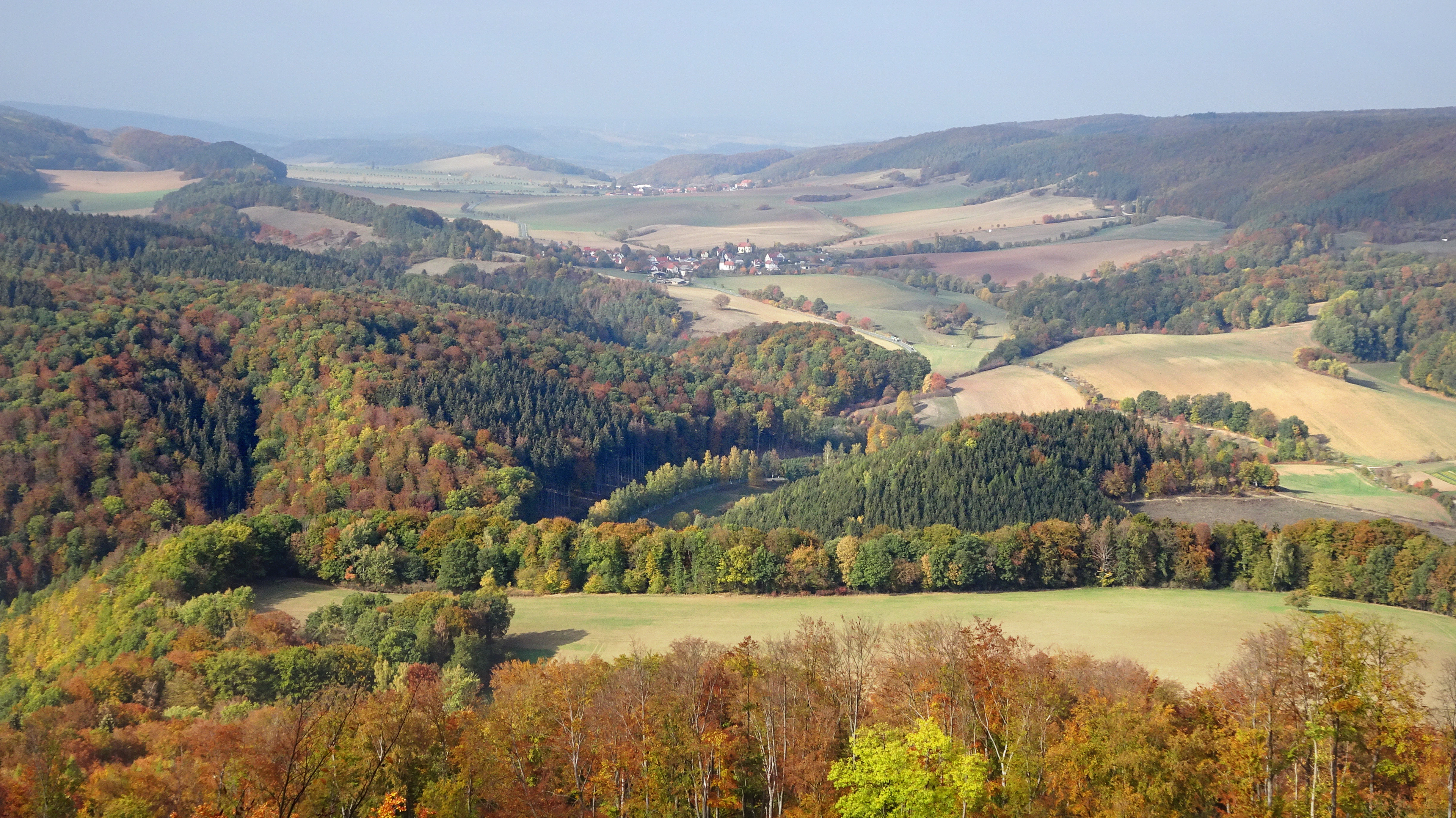 A wide, high-angle view of a rolling, forested valley in autumn with a small village nestled in the distance and colorful deciduous trees.