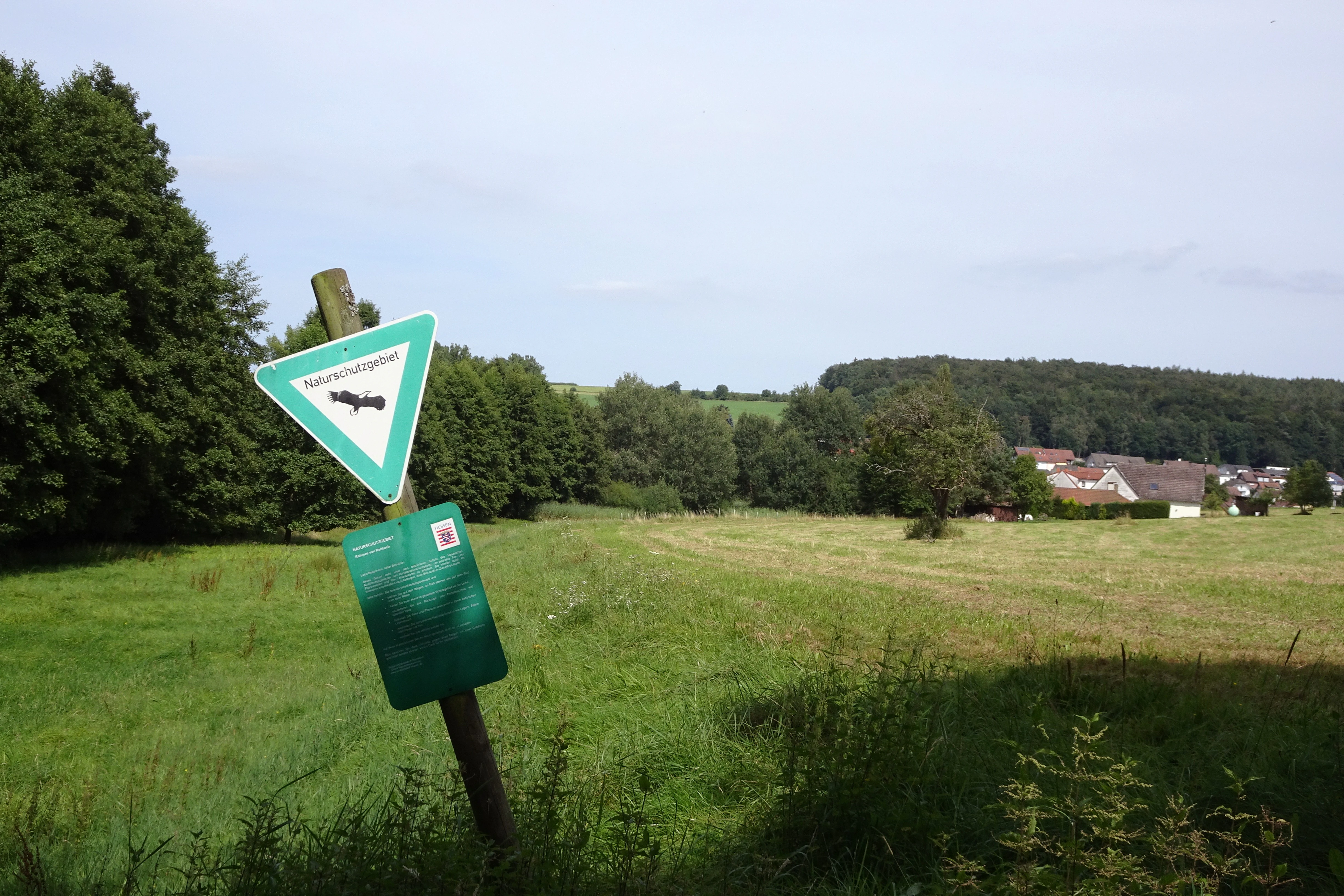 A wooden post with a green triangular sign featuring a bird silhouette and a rectangular information board stands in a grassy field. Trees, a forested hill, and houses are visible in the distance under a cloudy sky.