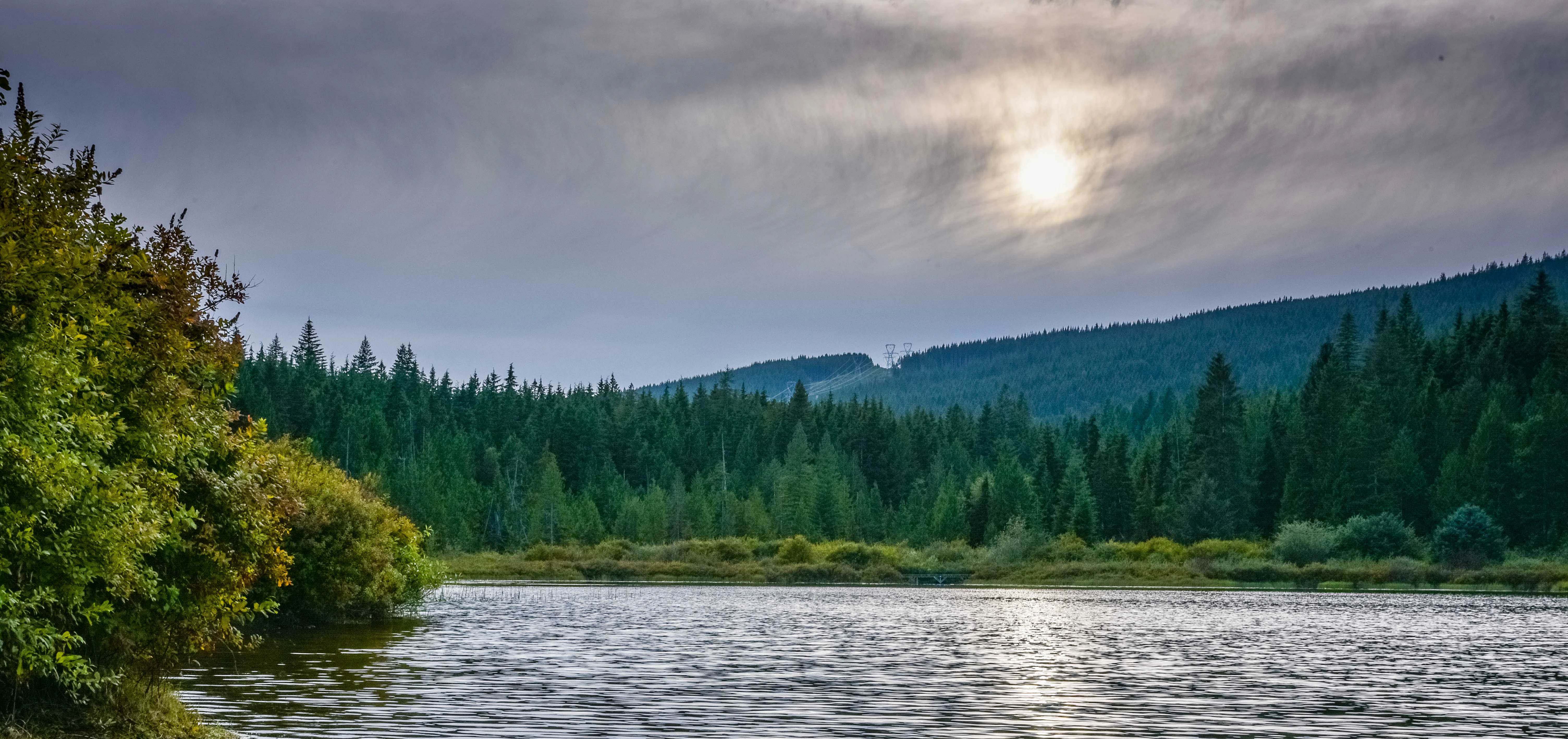 A calm lake surrounded by dense evergreen forest under a cloudy sky with the sun filtering through.