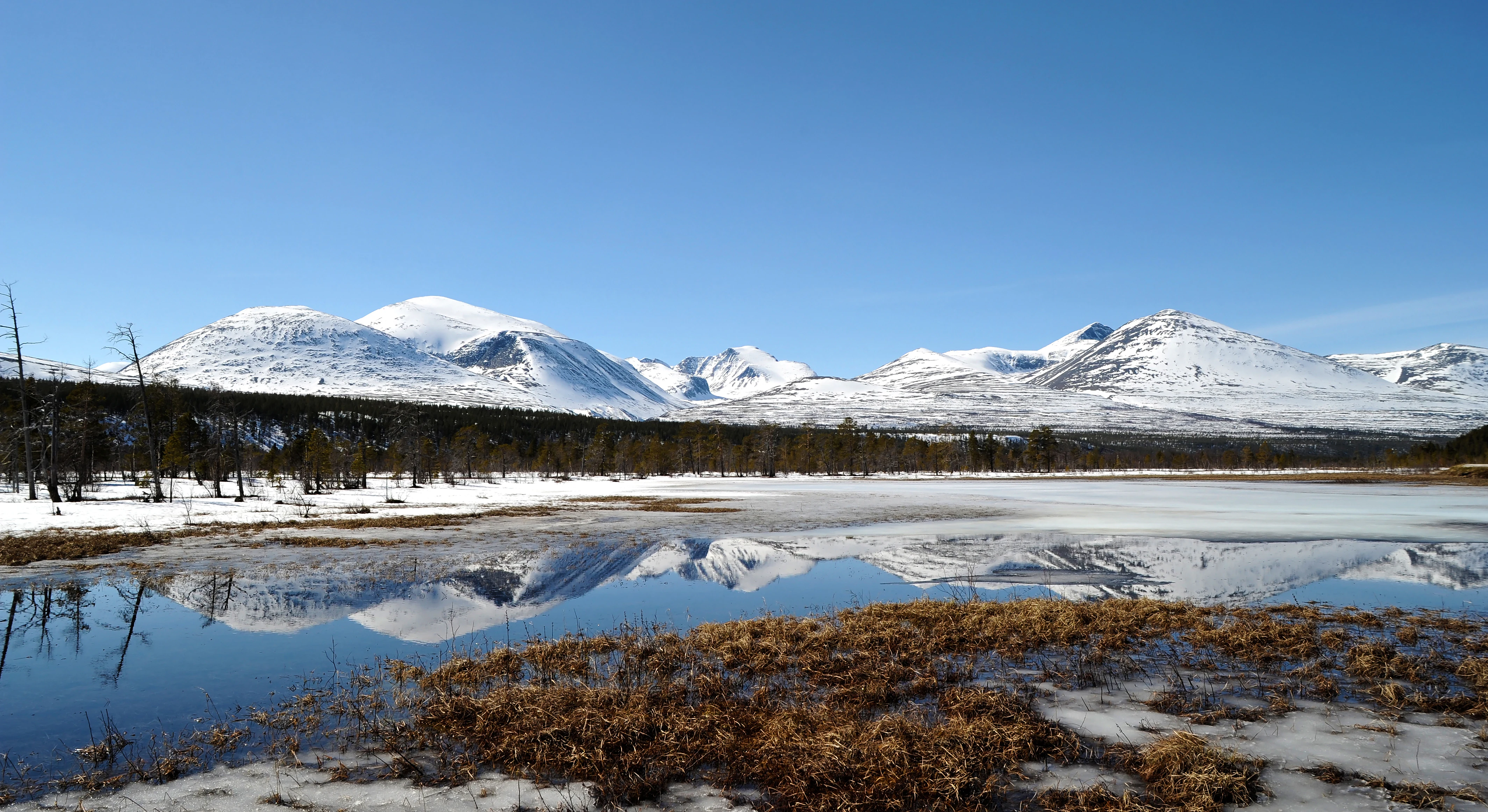 Snow-capped mountains reflecting in a calm lake with patches of snow on the shore and a clear blue sky