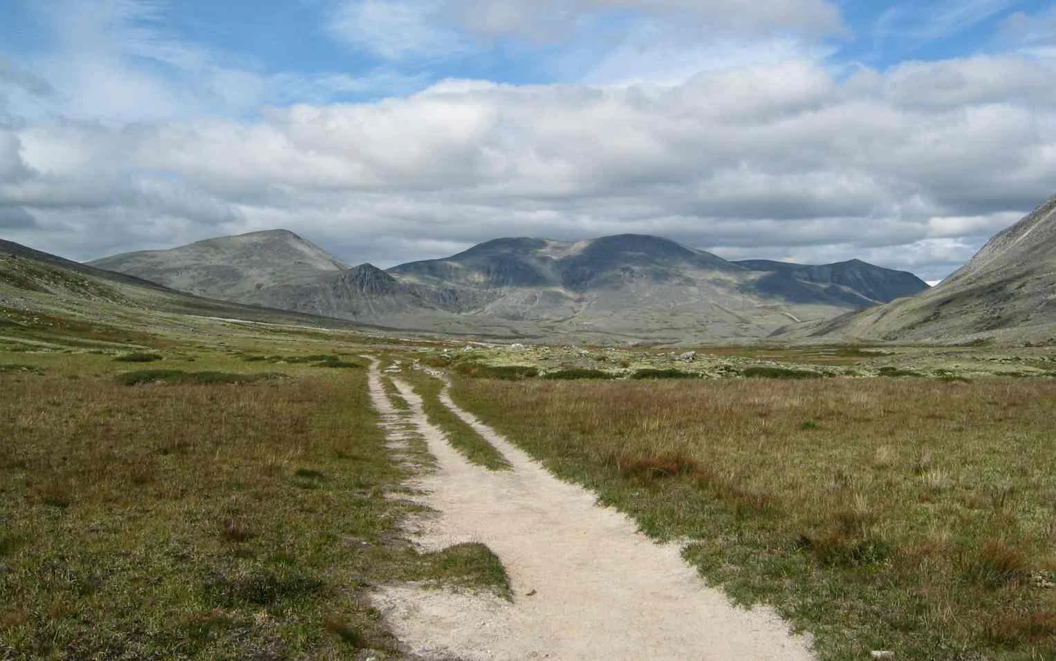 A dirt path through a grassy field with rolling hills and mountains under a partly cloudy sky.