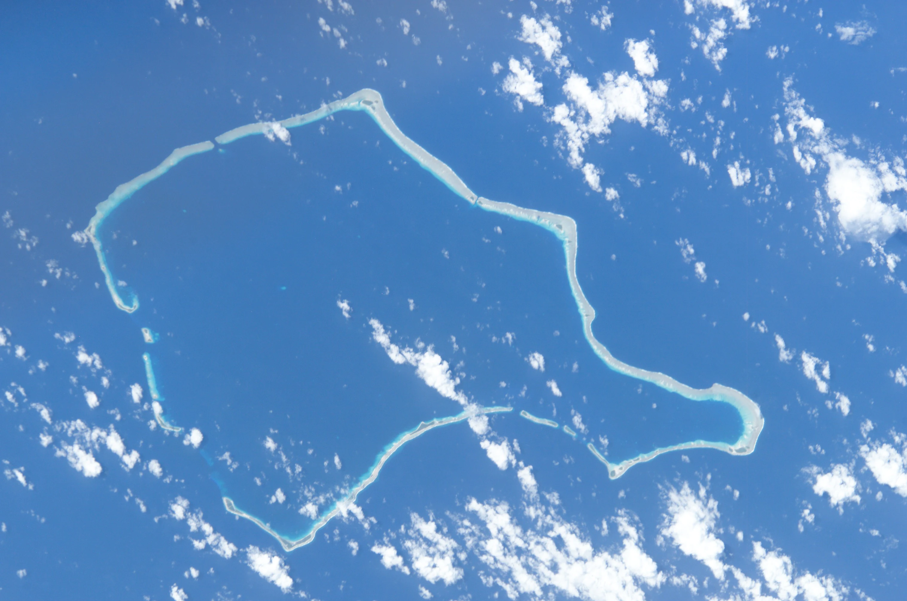 An aerial, high-angle view of Rongelap Atoll in the Pacific Ocean, featuring a jagged, circular ring of coral reefs and islands against a deep blue sea with scattered white clouds.