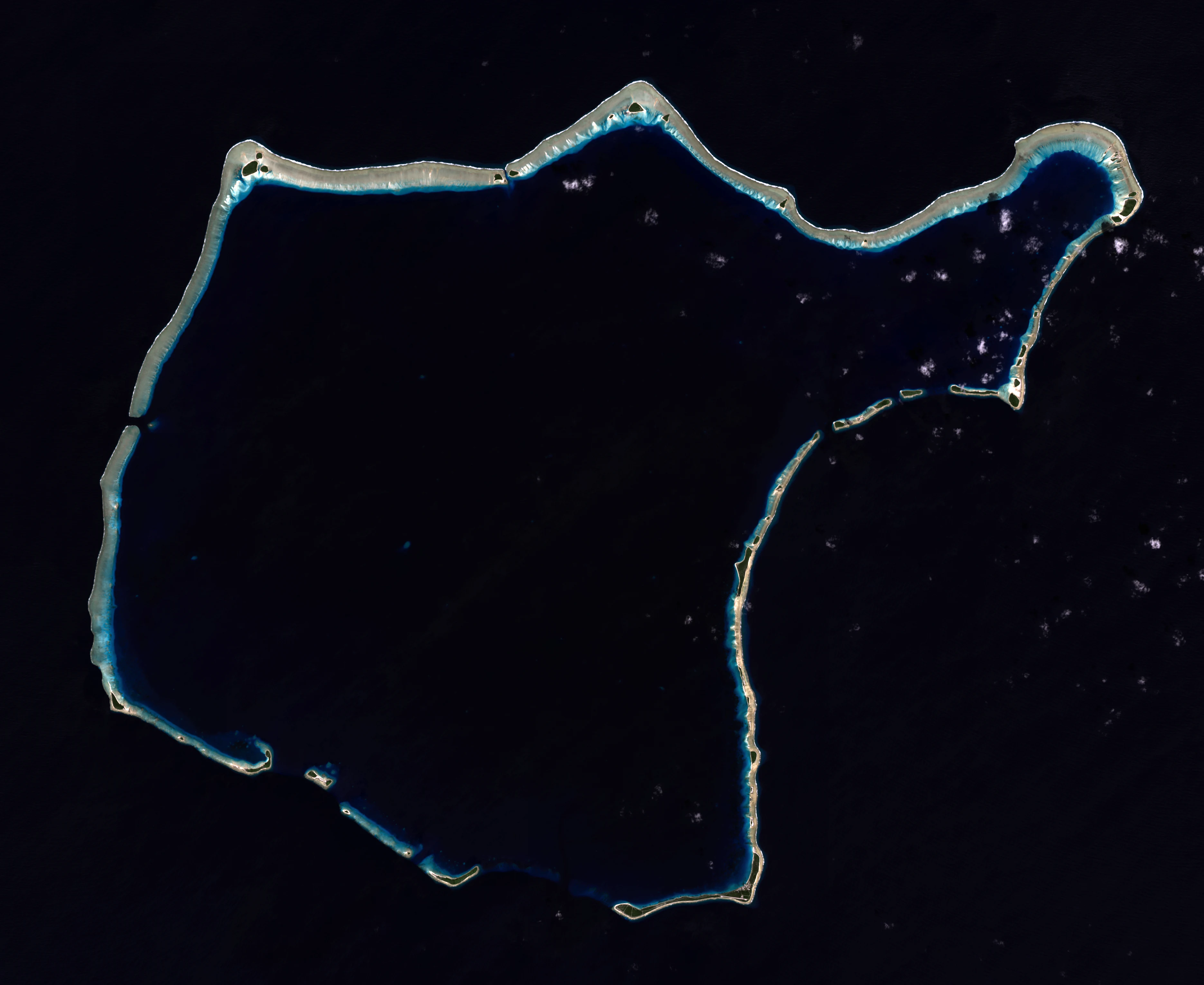 Satellite view of a coral atoll showing a ring of islands and reefs surrounding a central dark blue lagoon against a deep black ocean background.