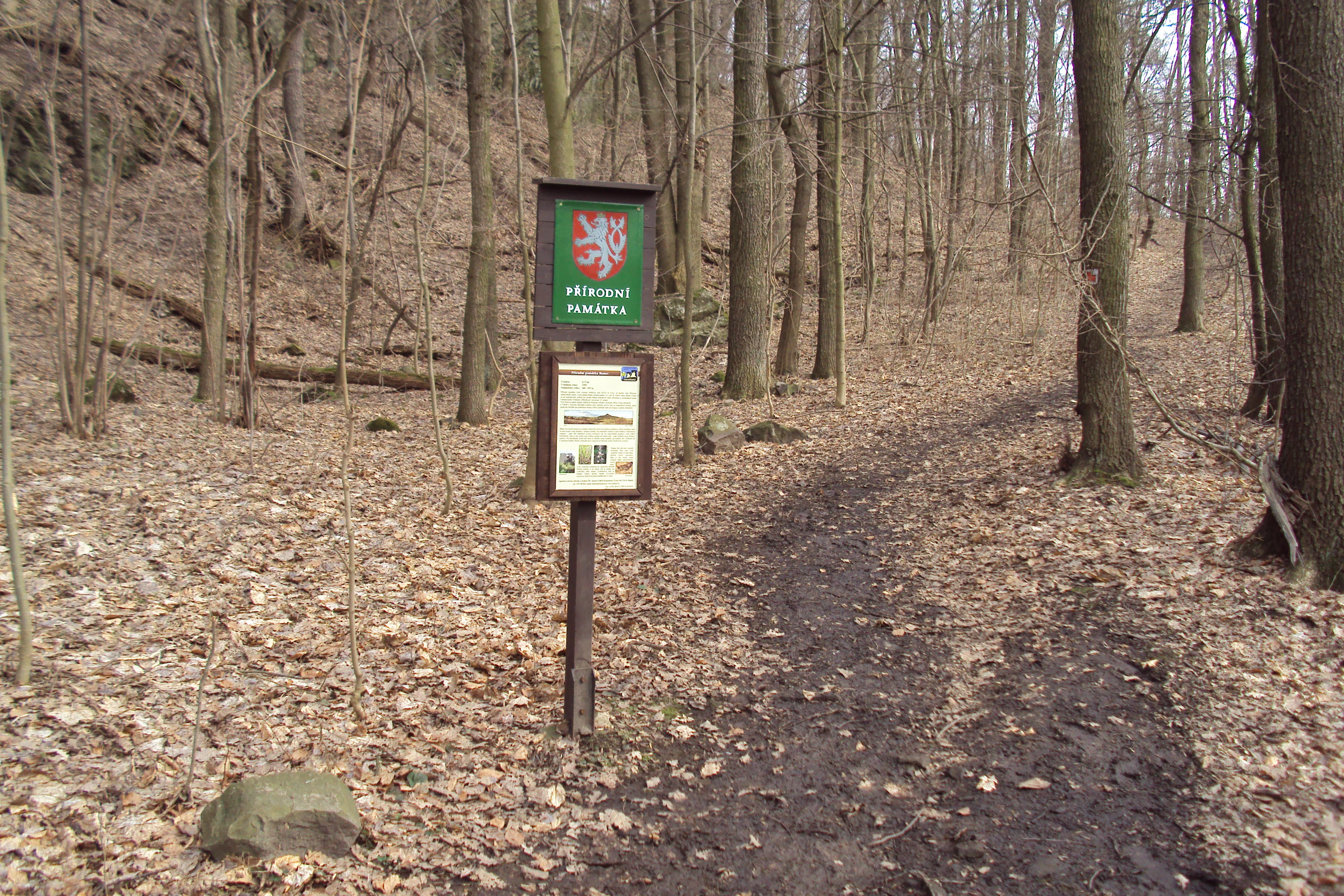 Un cartel de madera marrón de parque se alza sobre un poste junto a un sendero de tierra en un bosque caducifolio a finales de otoño o en invierno.