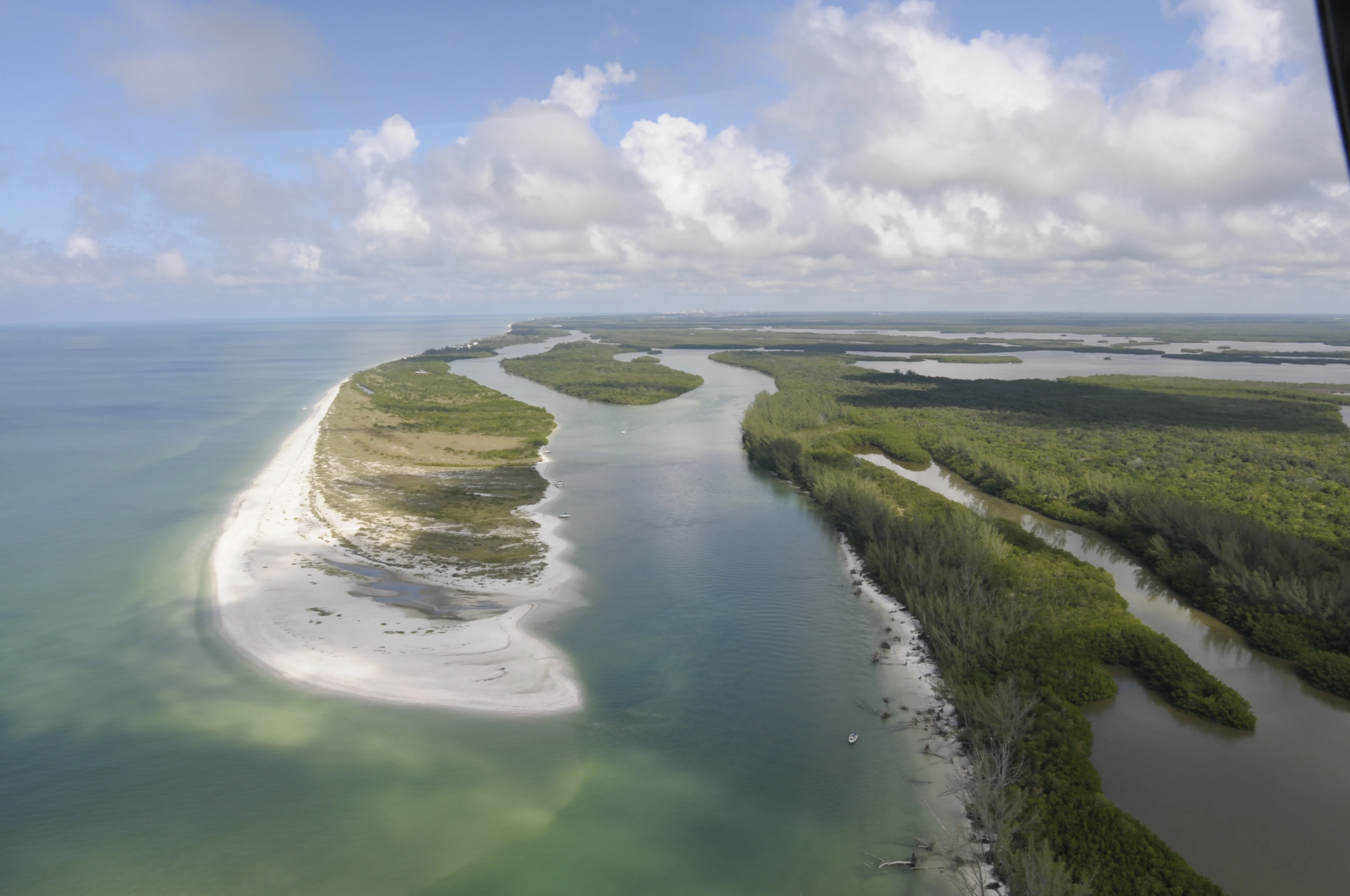 Aerial view looking over a sandy barrier island, tidal channels, and dense mangrove forests meeting the open ocean under a partly cloudy sky.