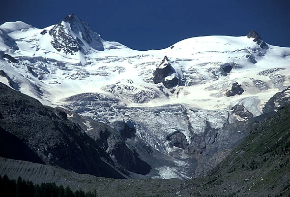 A wide angle view of the massive snow and ice covered Roseggletscher surrounded by dark rocky alpine mountain peaks under a clear blue sky.
