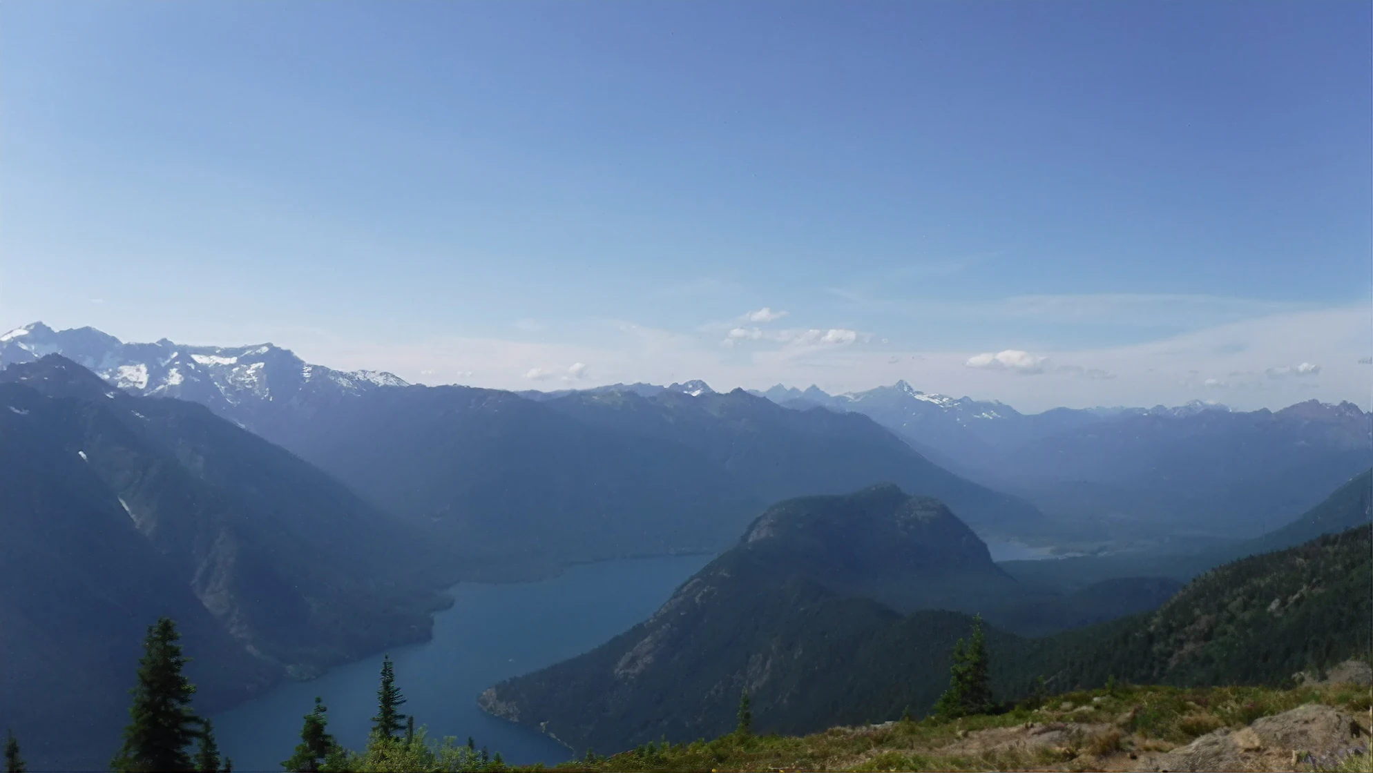 A wide, elevated mountain landscape view looking down onto the deep blue waters of Ross Lake, flanked by forested ridges and distant snow-capped peaks under a clear sky.