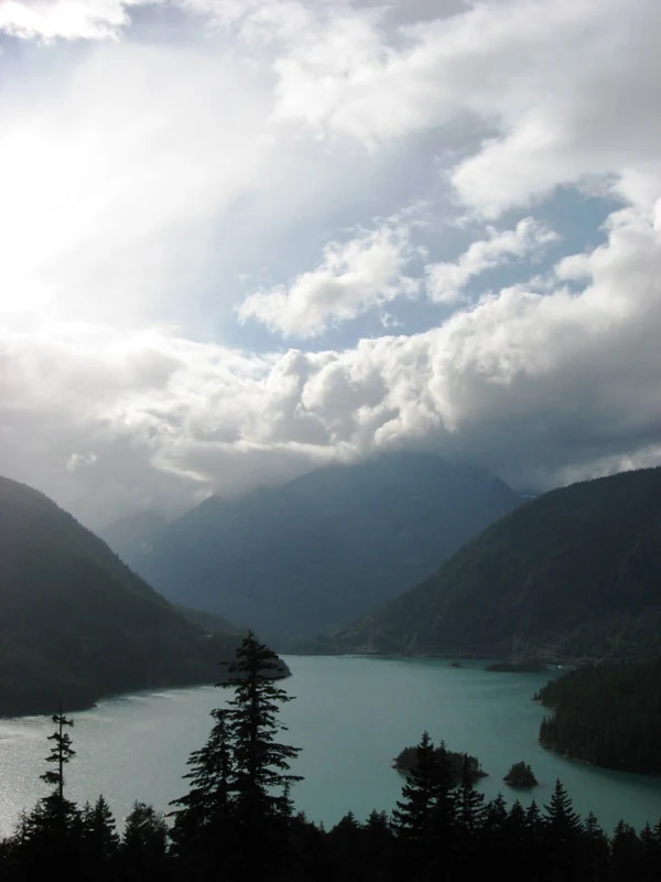 A high-angle view looking over the turquoise water of Diablo Lake, surrounded by dark forested mountains under a cloudy sky.