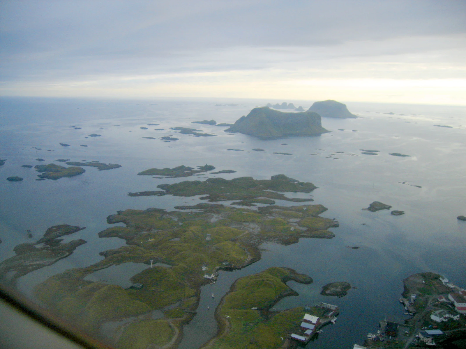 An aerial shot looking over the Røst archipelago in Norway, with many small grassy islands scattered across blue coastal waters under a cloudy sky.
