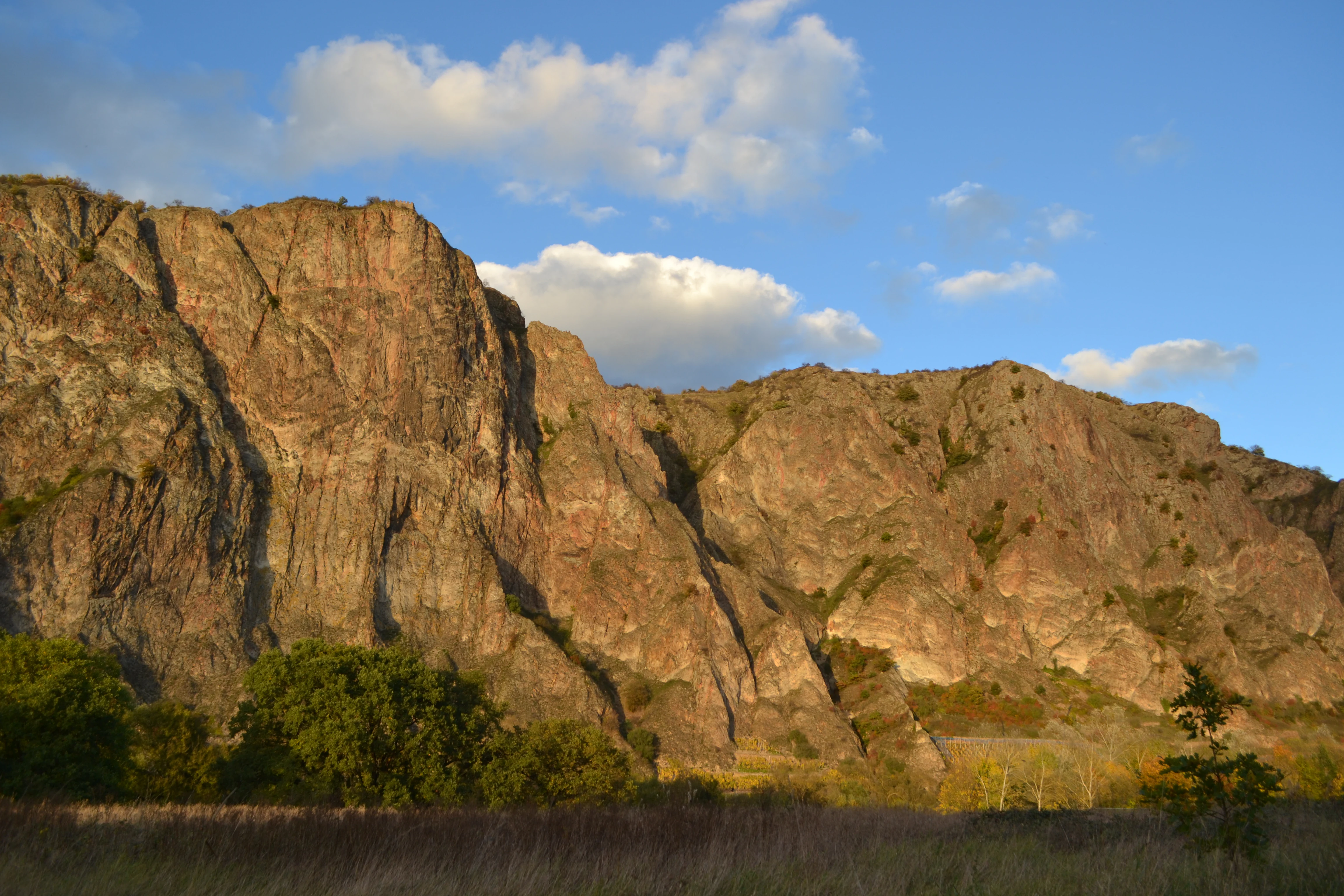 A wide shot of a massive, steep, rocky mountain cliff face rising above a landscape of trees and grasses under a clear blue sky with white clouds.