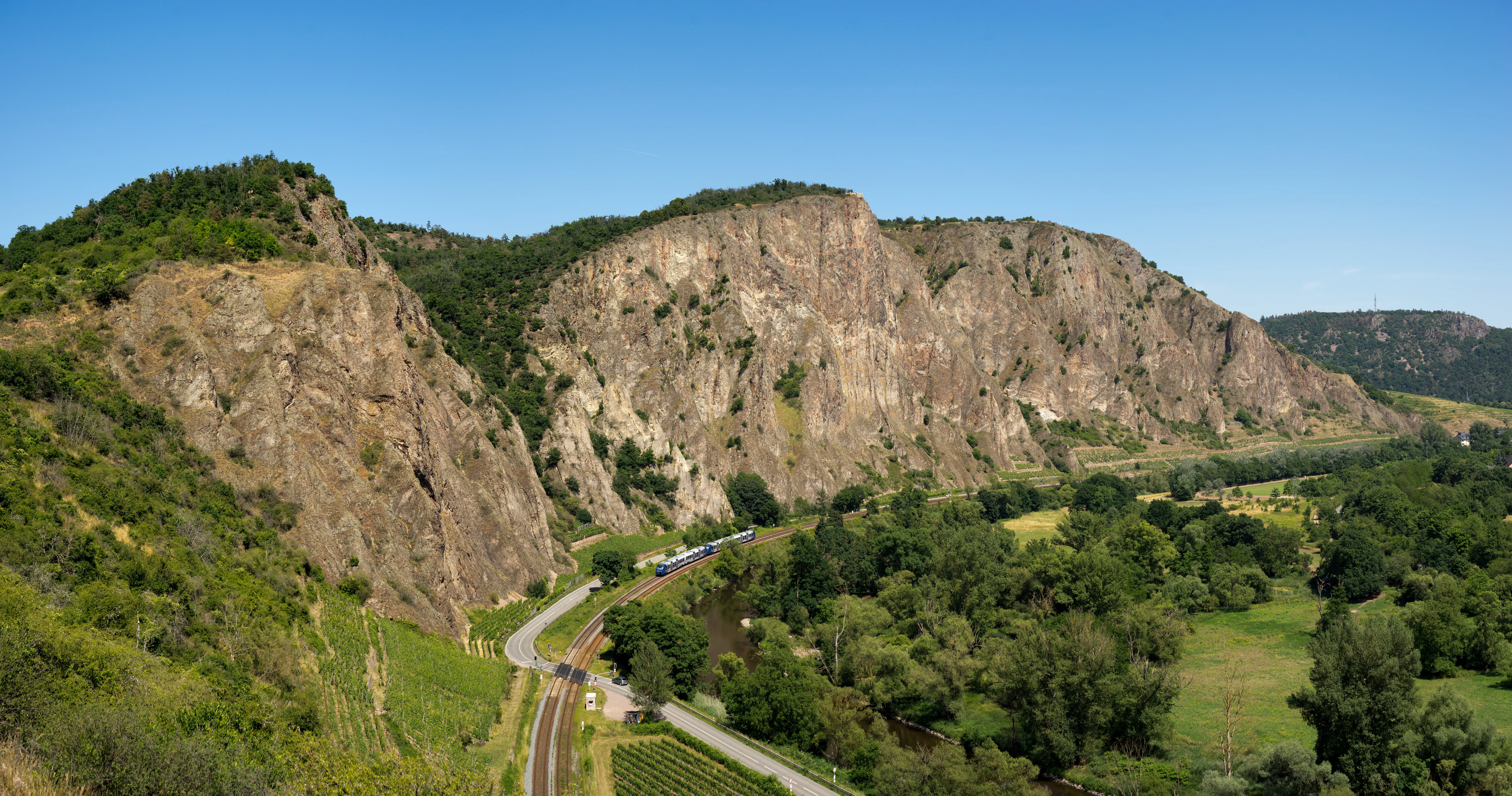 A wide view of the tall, vertical, reddish-brown rock walls of the Rotenfels mountain massif, with a railway track and lush green vegetation in the foreground.