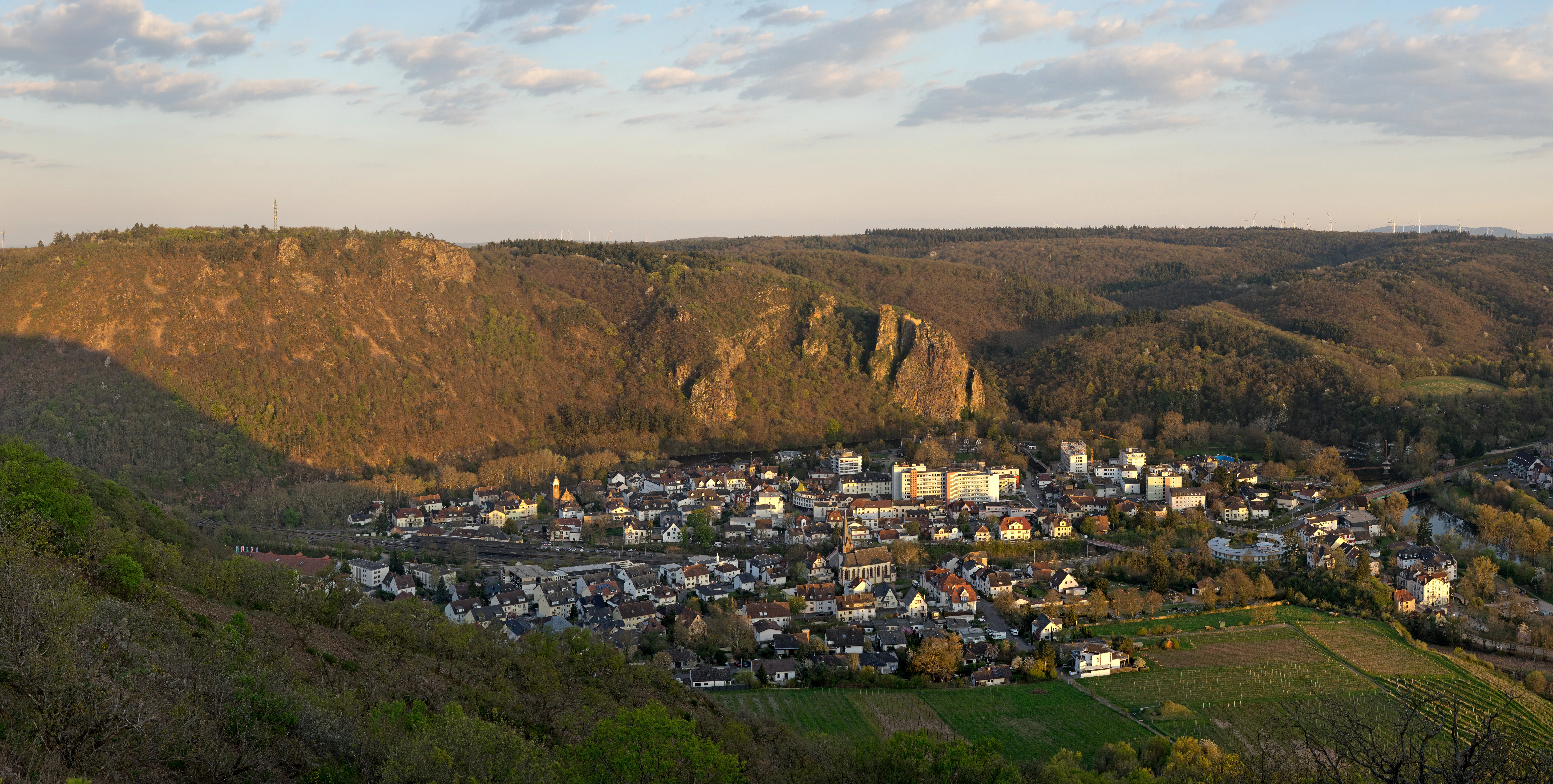 A high-angle panoramic view looking down from the rocky heights of Rotenfels onto a small town situated in a valley, with rolling forested hills in the background during golden hour.