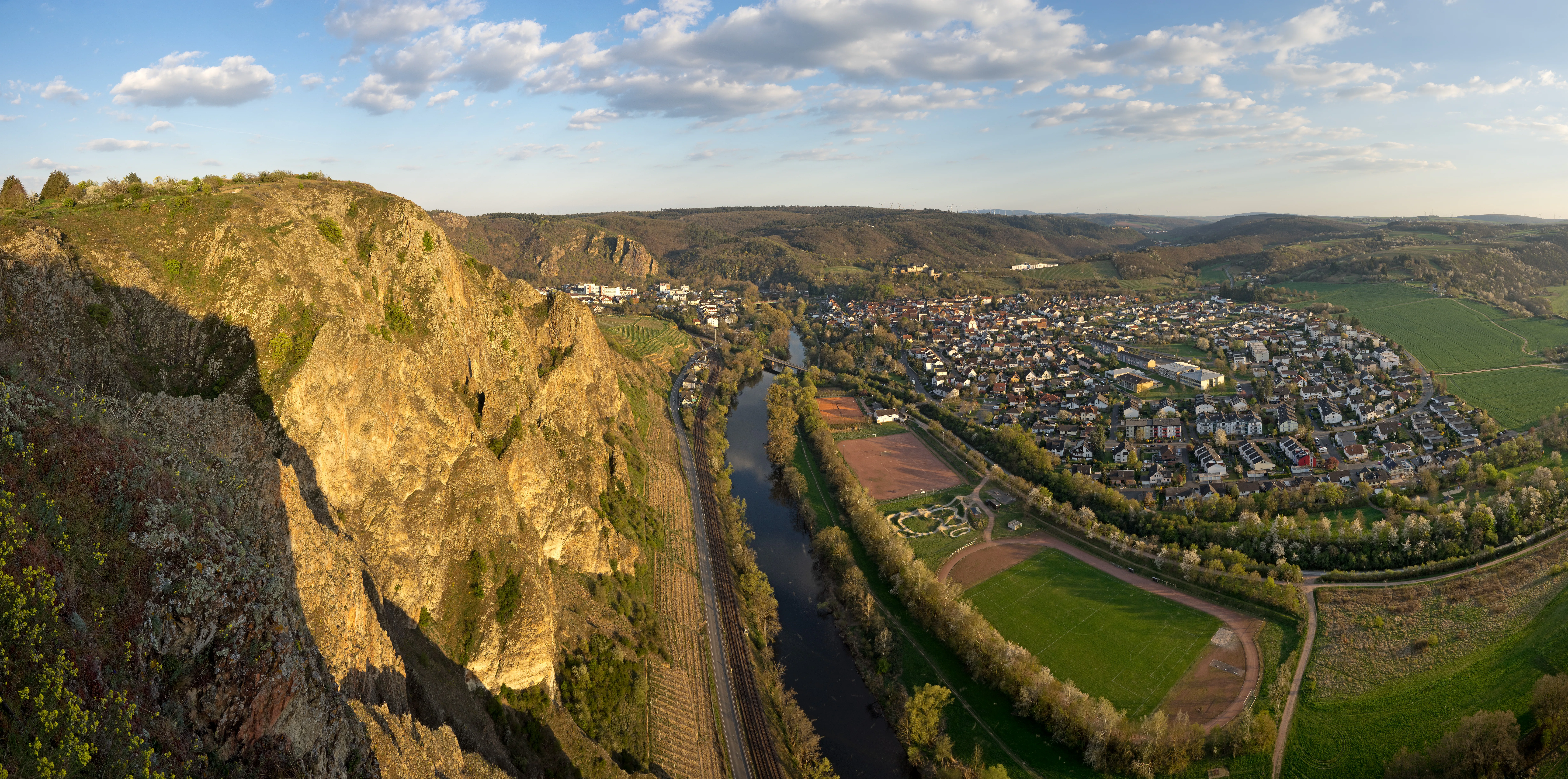 Wide panoramic view from the Bastei viewpoint at Rotenfels, showing a high, sheer vertical rock face on the left, a river flowing through the valley below, and a town nestled along the riverbank.
