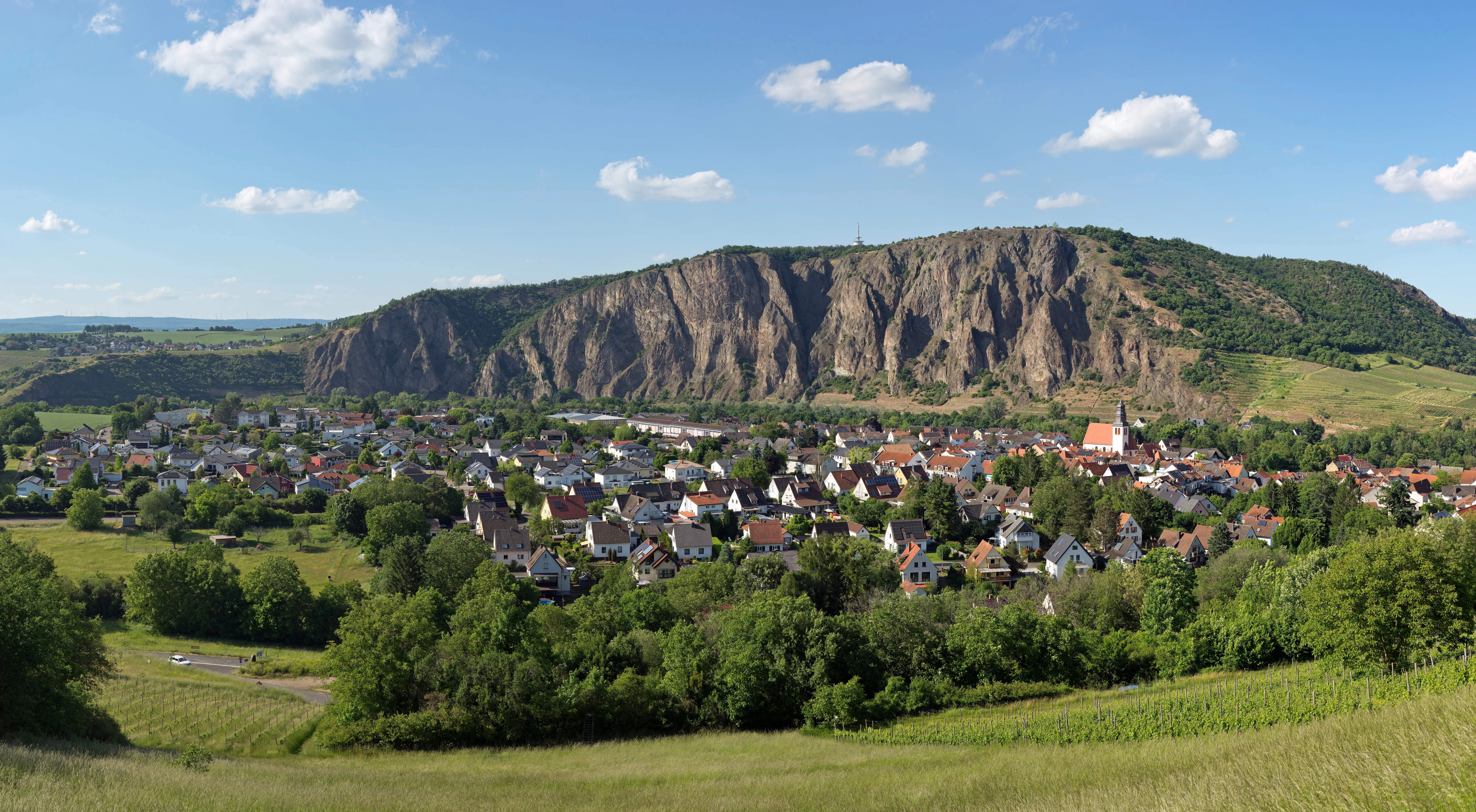 A wide landscape view of the steep, jagged, and rocky Rotenfels massif towering over a small town with red-roofed buildings, seen under a sunny sky.