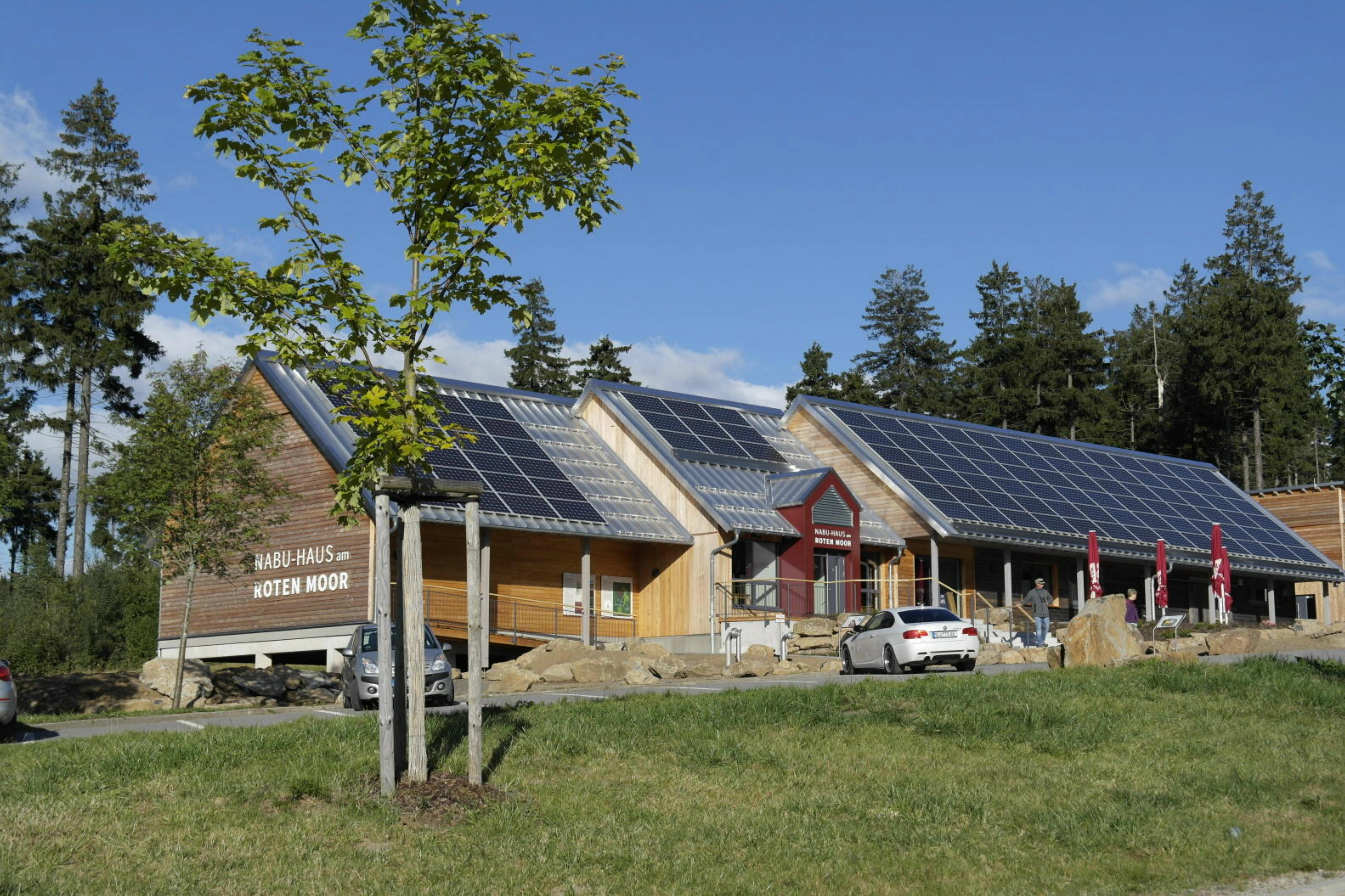 A modern wooden visitor center building with solar panels on its slanted roof, surrounded by trees and a grassy foreground.