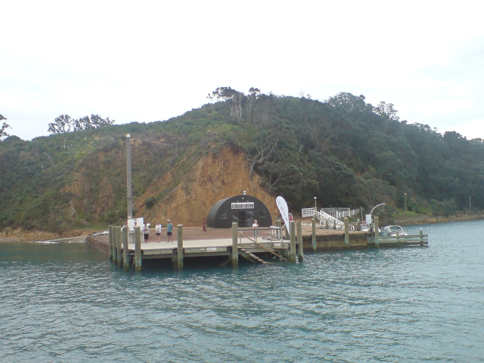 A view from the water looking at a wooden ferry pier on Rotoroa Island, featuring a dark rounded shelter building at the base of a grassy, forested hill.
