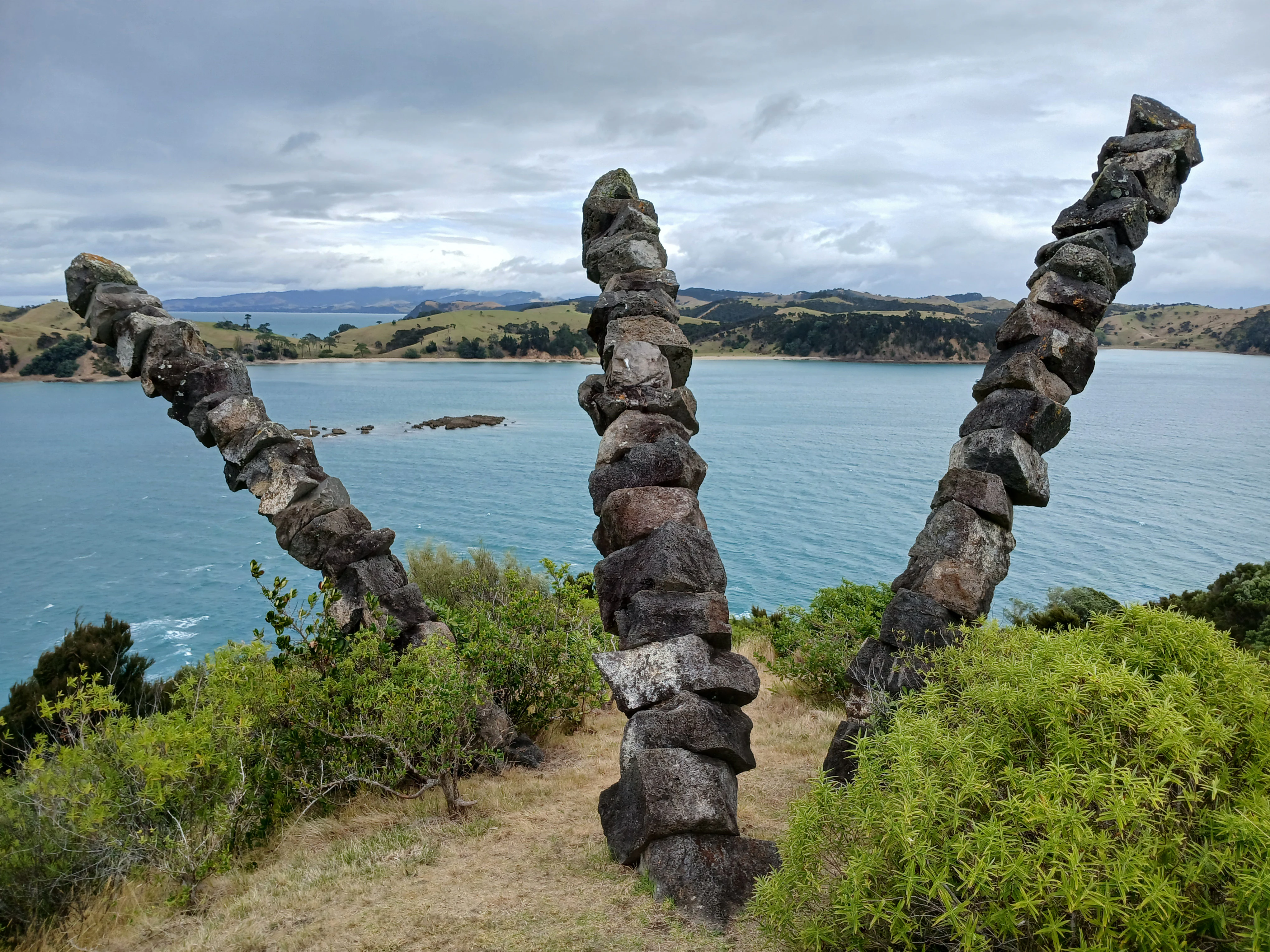 Three tall, slender stacks of dark volcanic rocks, resembling abstract towers, stand on a grassy ridge overlooking a wide blue bay with rolling hills in the background.