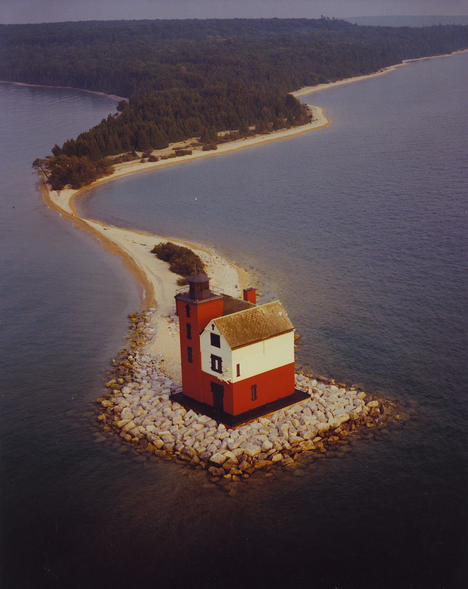 An aerial view of the red and white Round Island Lighthouse situated on a rocky foundation at the end of a long, curving sandy beach and forested island.