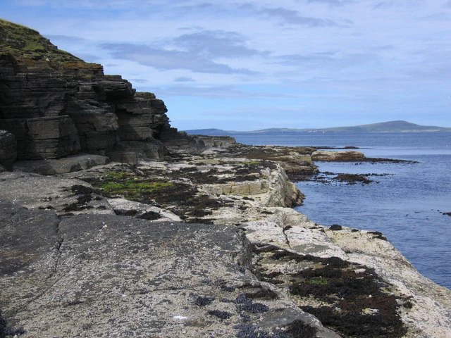Rugged, layered rocky cliffs on the left sloping down to a low, flat shoreline bordering the sea, with distant islands visible across the water under a cloudy sky.