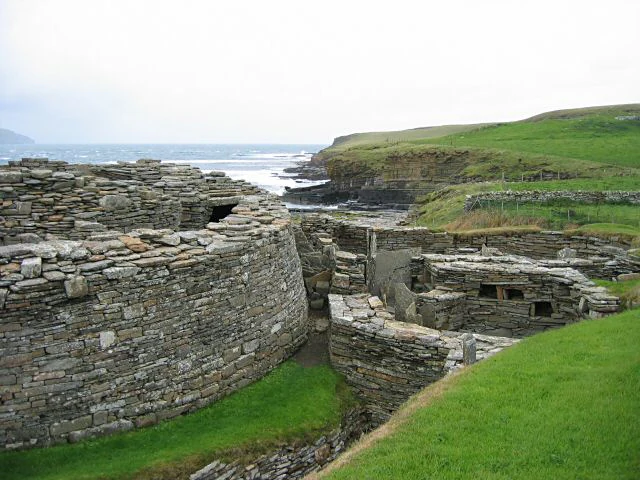 Ancient stone structure of Midhowe Broch showing circular walls and interior chambers next to the grassy coastline.