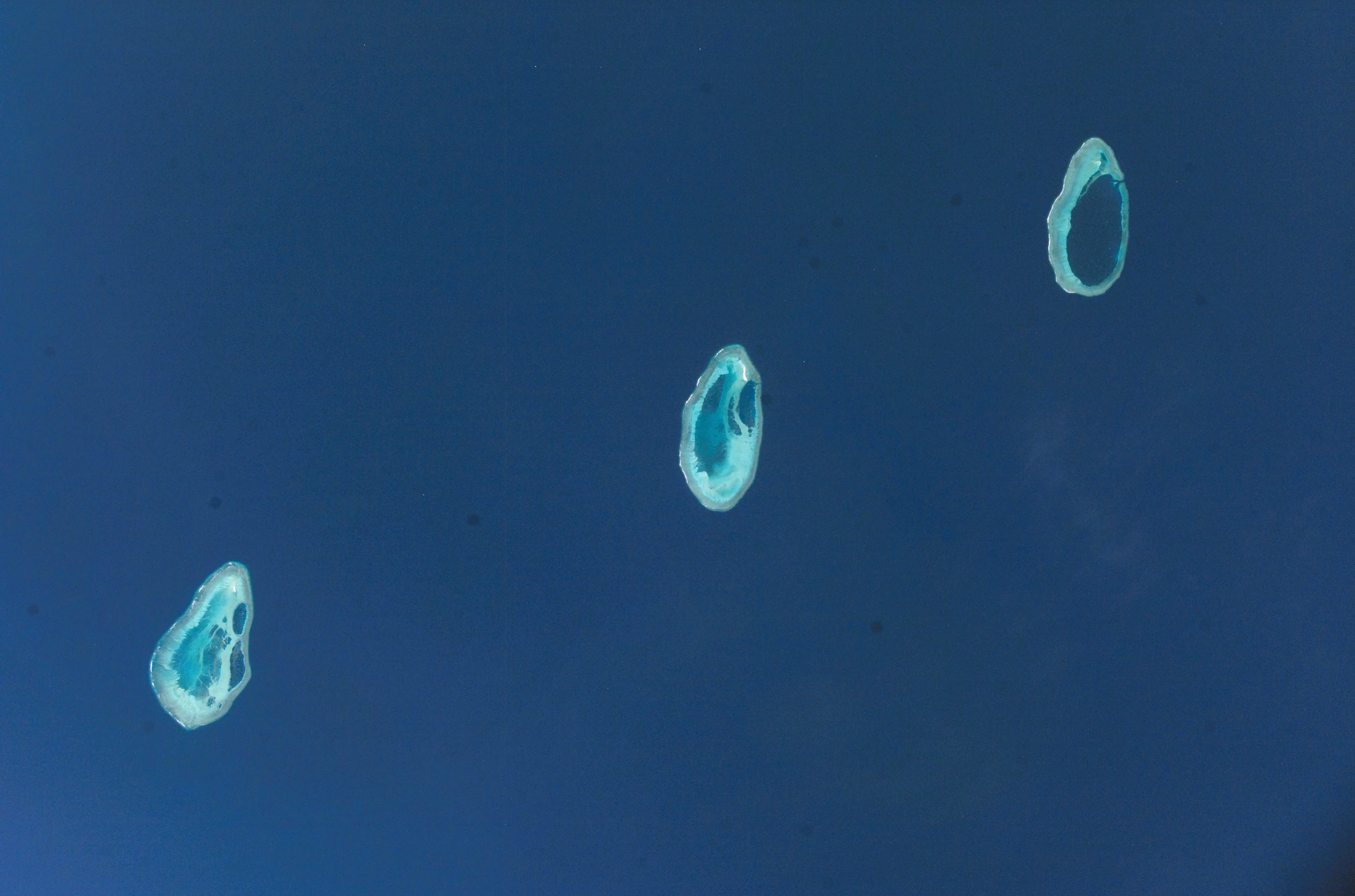 Satellite aerial view of three oval shaped coral atolls situated in the deep blue waters of the Indian Ocean.