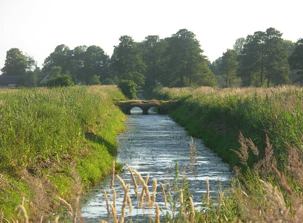 A narrow, straight canal cuts through green, grassy meadows leading toward a small stone bridge in the distance under a bright sky.