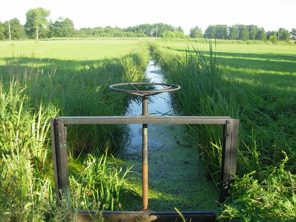 A close-up view of a metal manual sluice gate control wheel installed over a narrow, grassy drainage canal in a meadow landscape.