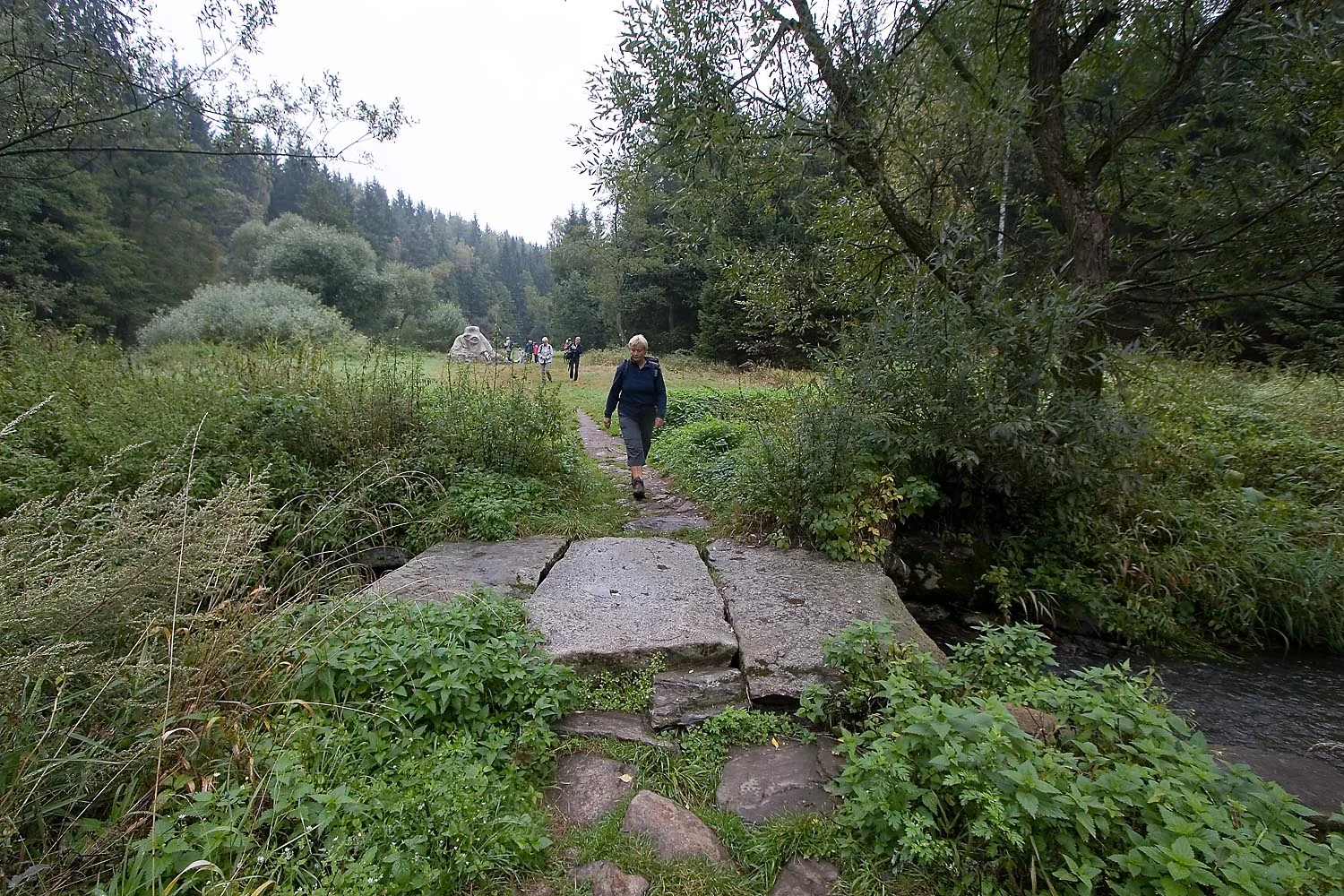 A hiker walking across a stone slab bridge over a small river in a forested landscape.