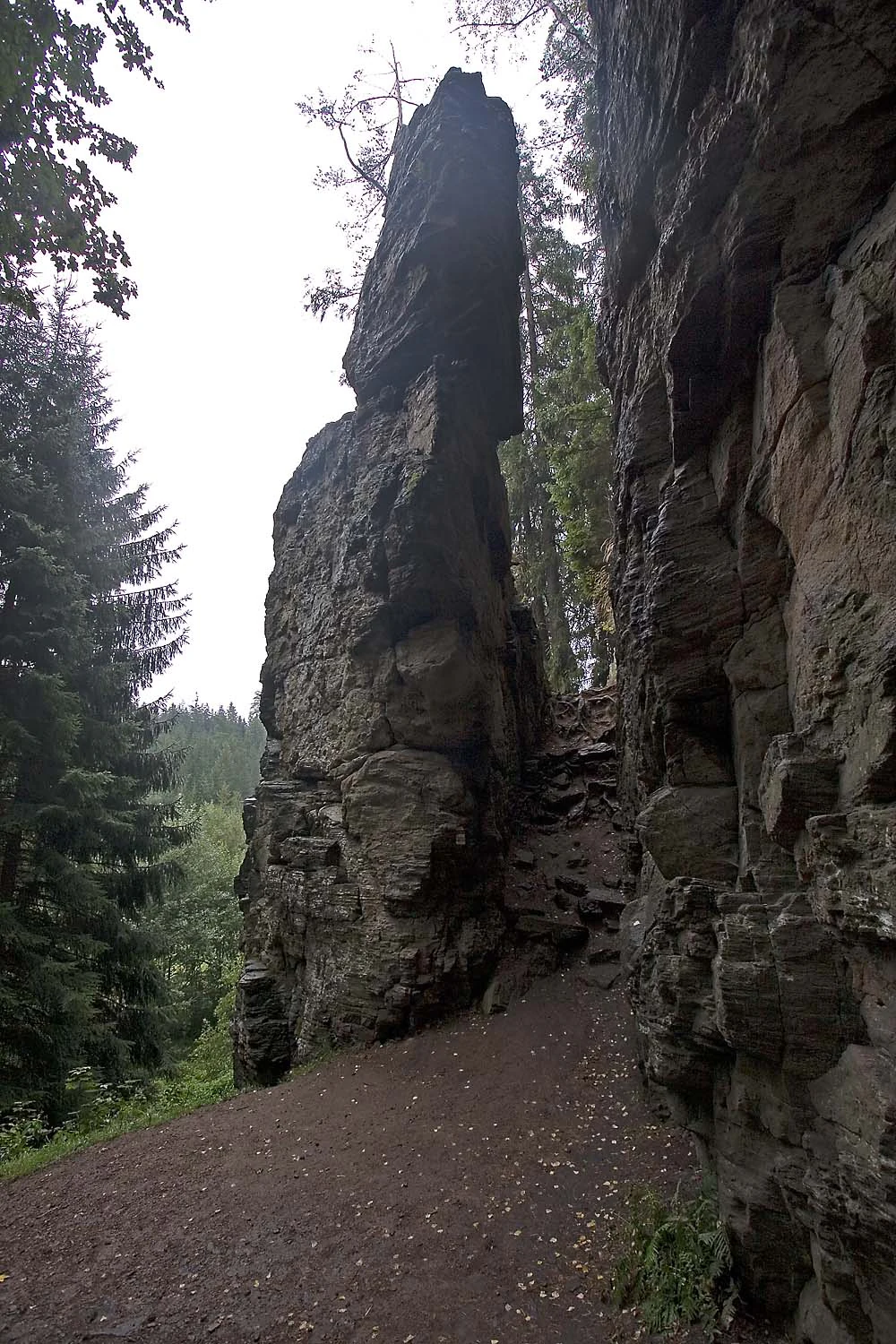 A vertical view of the tall gneiss rock formation at Rozštípená skála, showing a distinct split between two rock faces with a dirt path in the foreground.