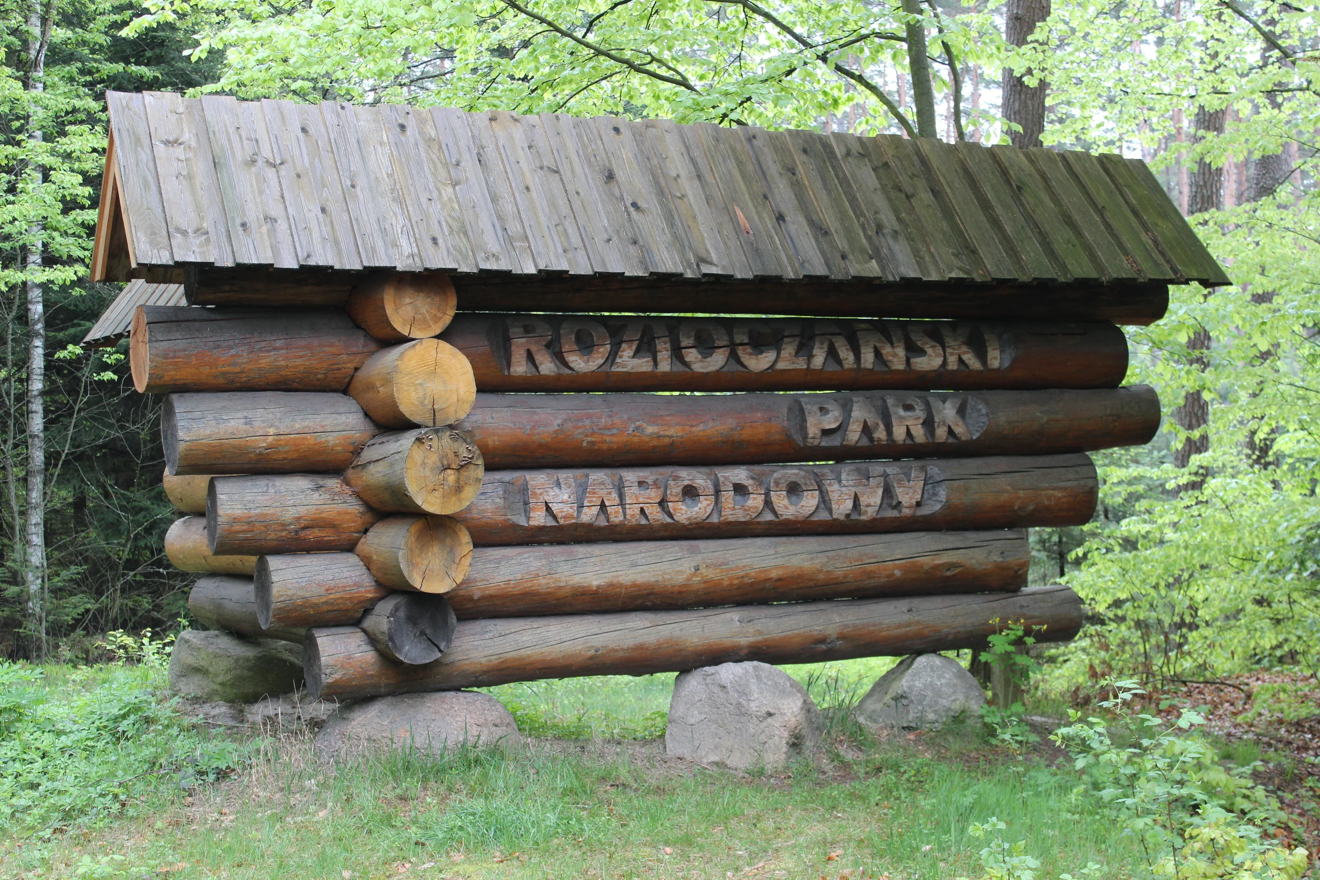 Wooden log sign with metal lettering 'ROZTOCZAŃSKI PARK NARODOWY' set in a forested area with green grass and trees